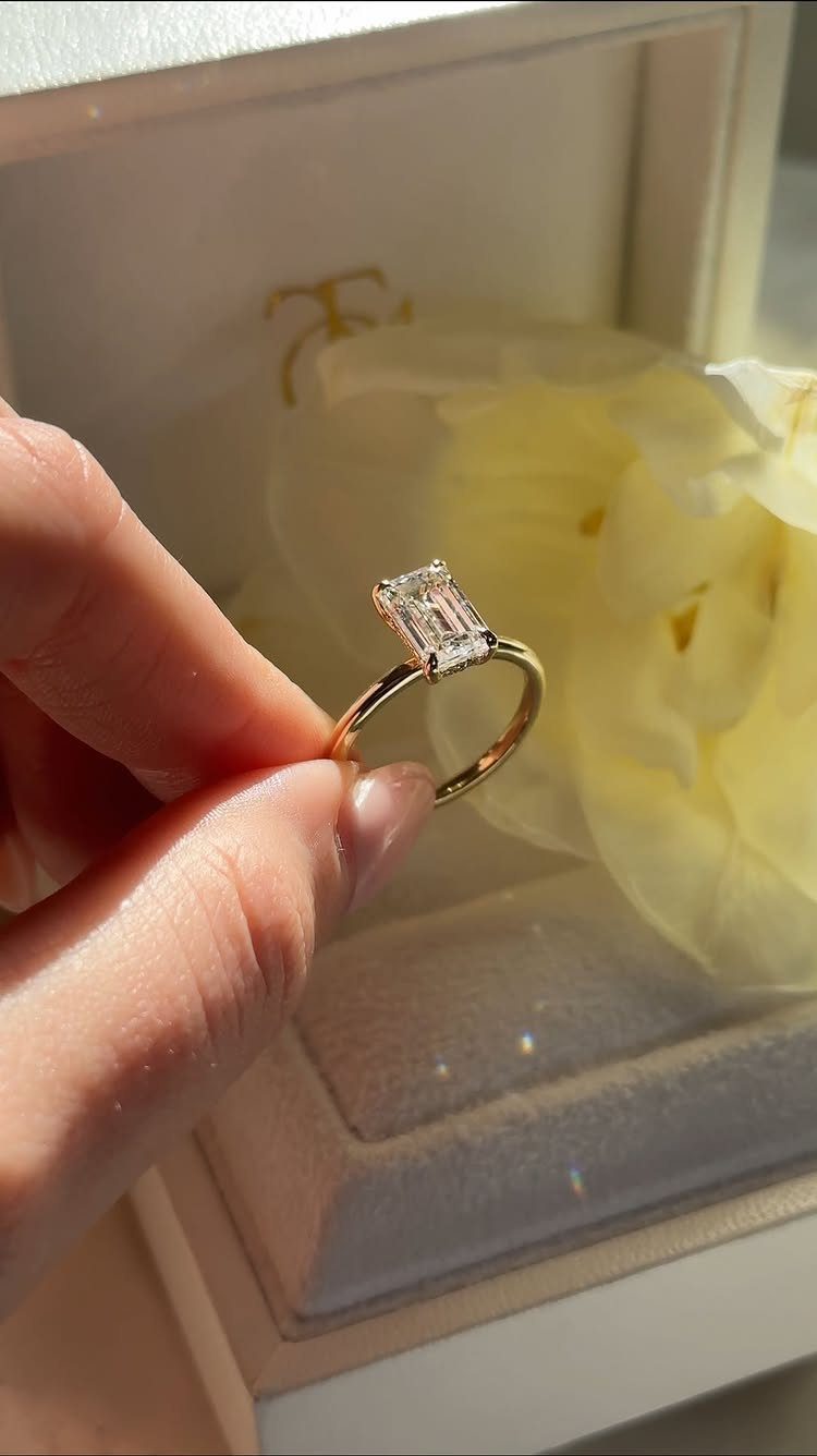 Gold engagement ring with a rectangular emerald-cut diamond held against a yellow flower.