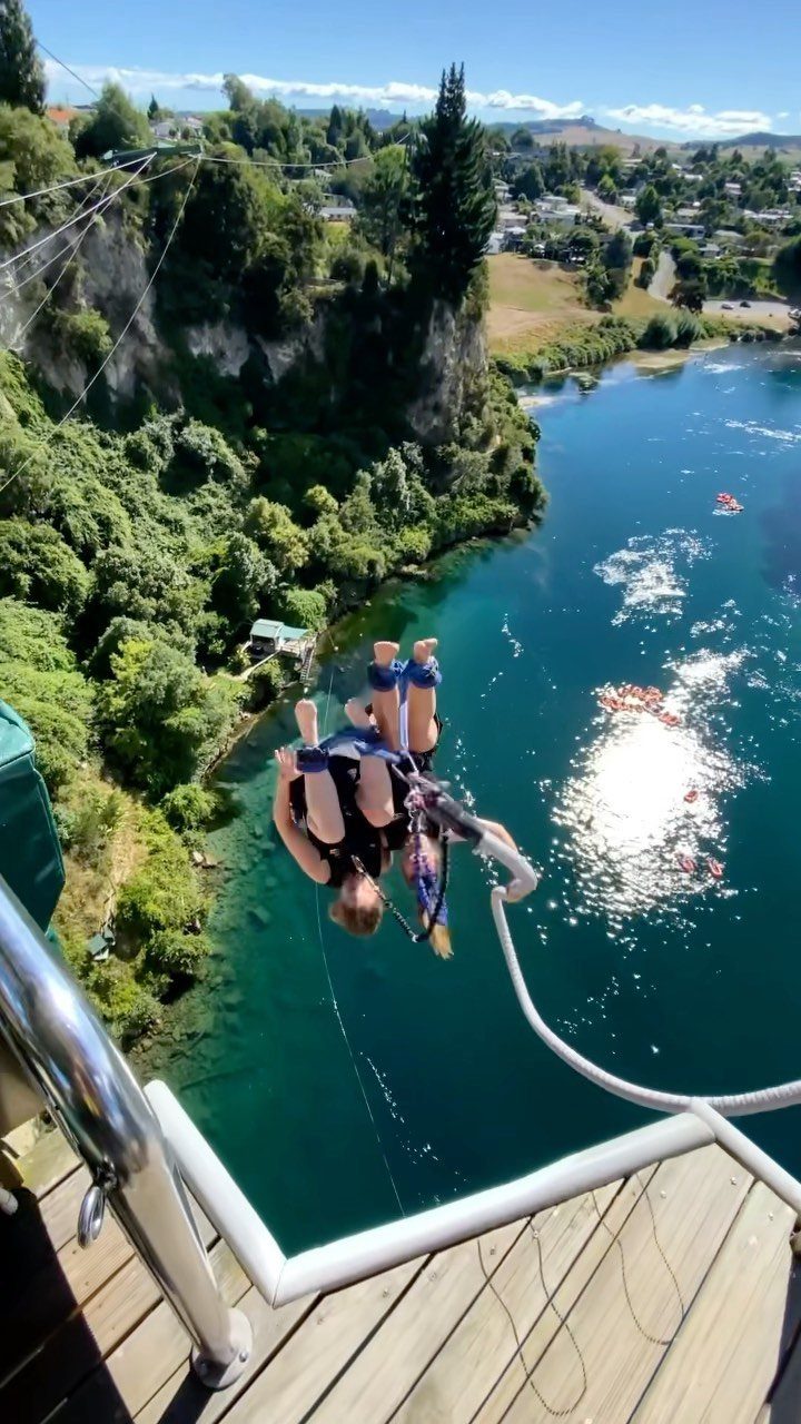 Person bungee jumping from a platform over a blue river and green cliffs.
