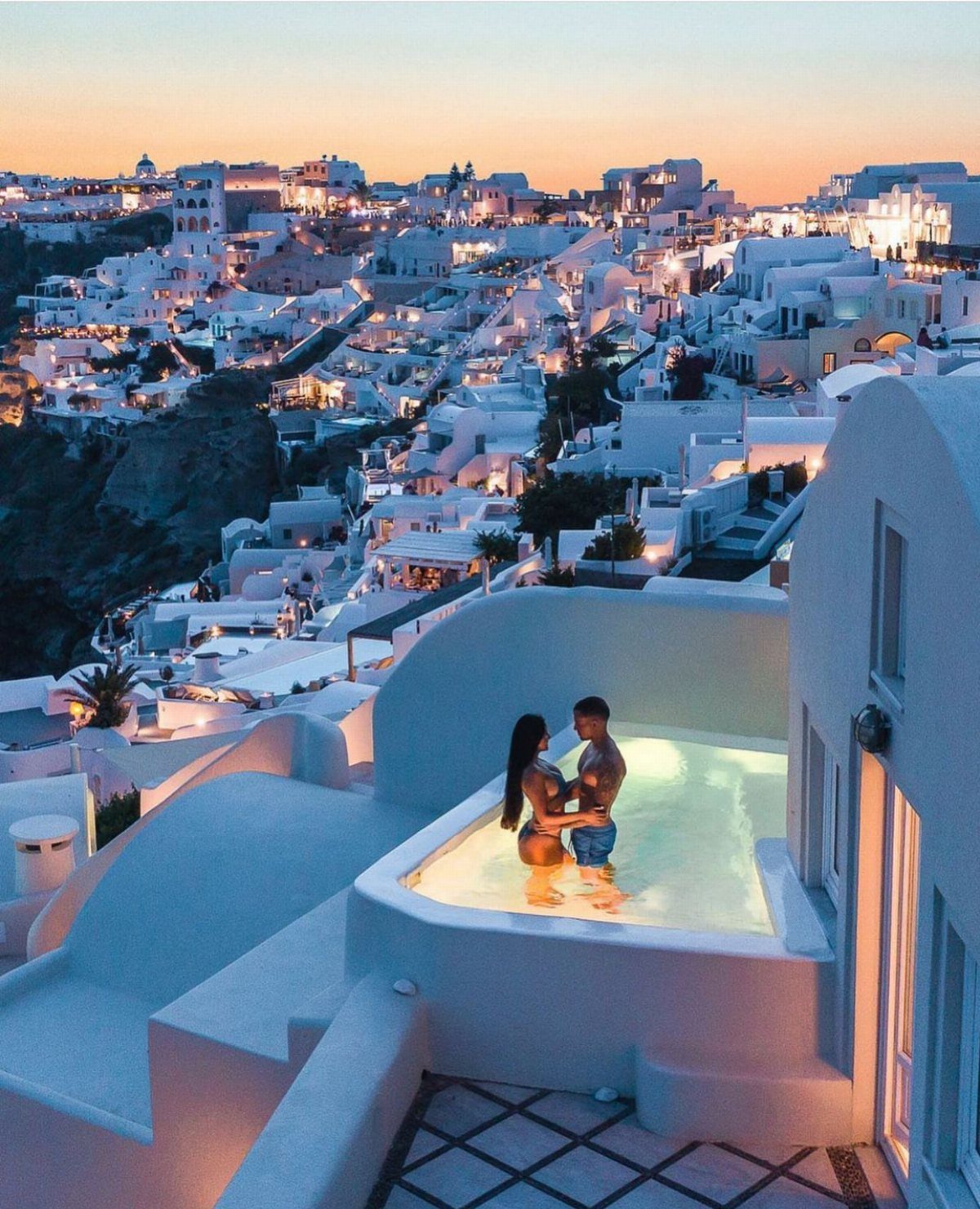 Couple in a glowing infinity pool overlooking the white buildings of Santorini, Greece.
