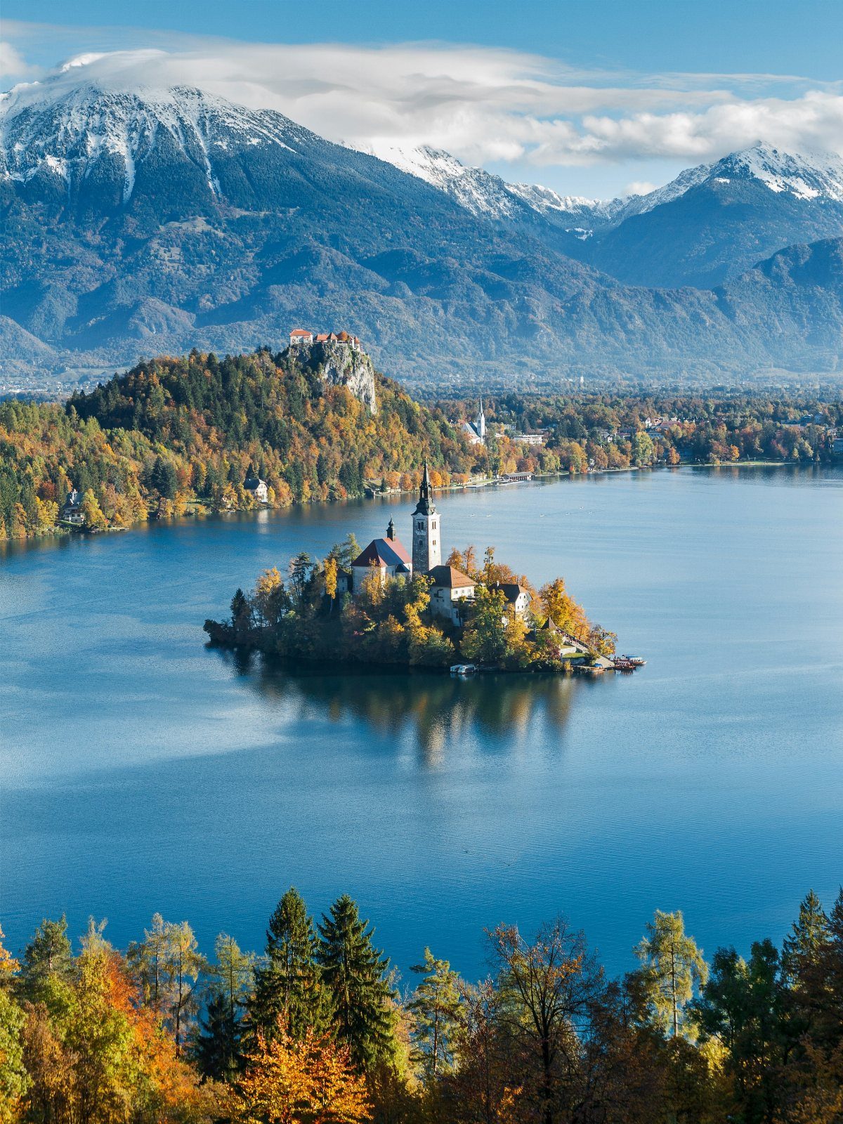 Lake Bled island with a church surrounded by autumn trees and snowy mountains.