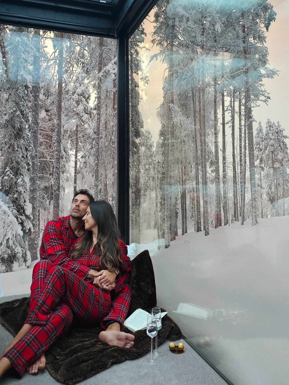 Couple in red plaid pajamas relaxing in a glass cabin overlooking snowy Finnish forests.