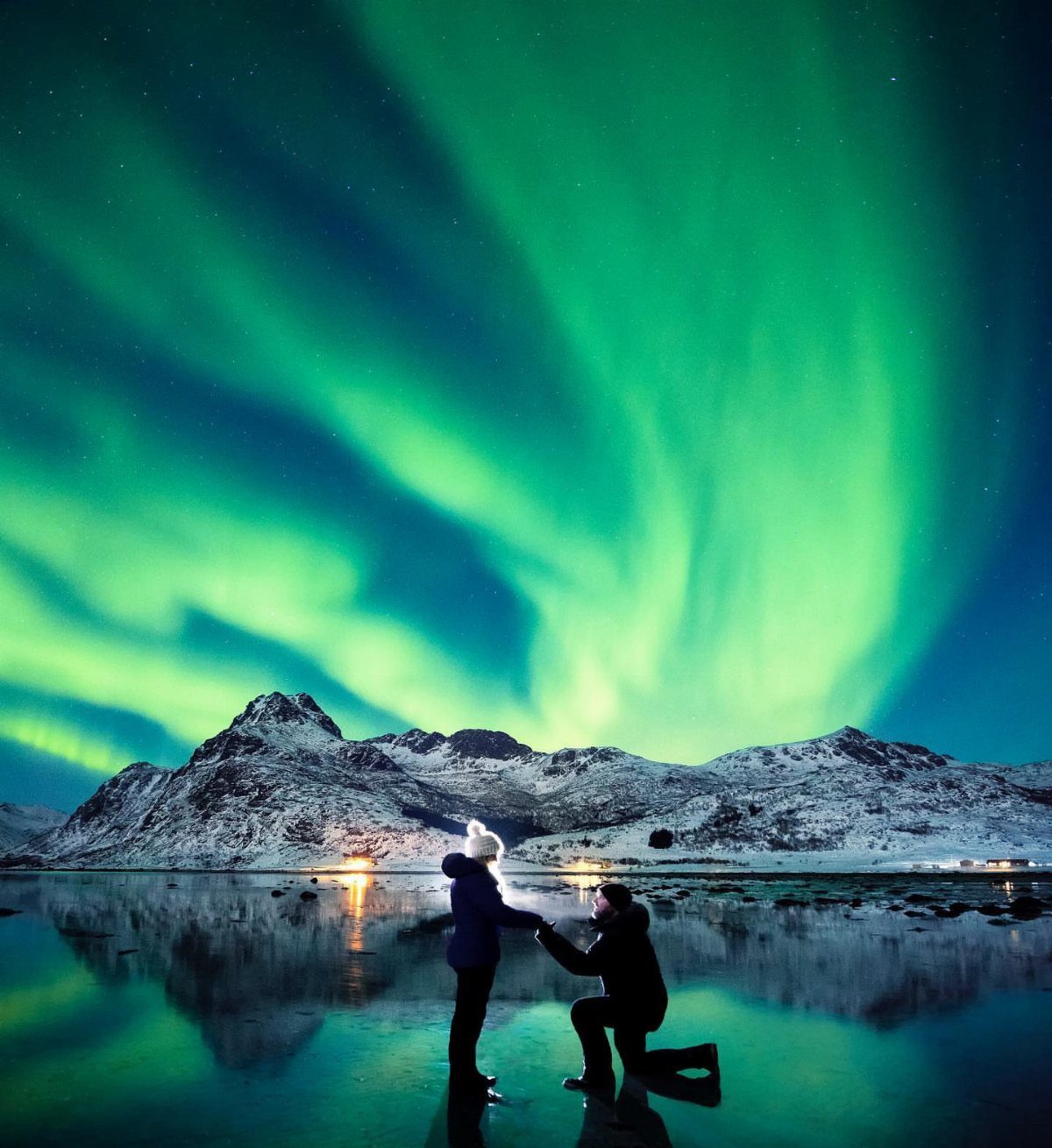 Couple proposing under a bright green aurora borealis with snowy mountains and water.