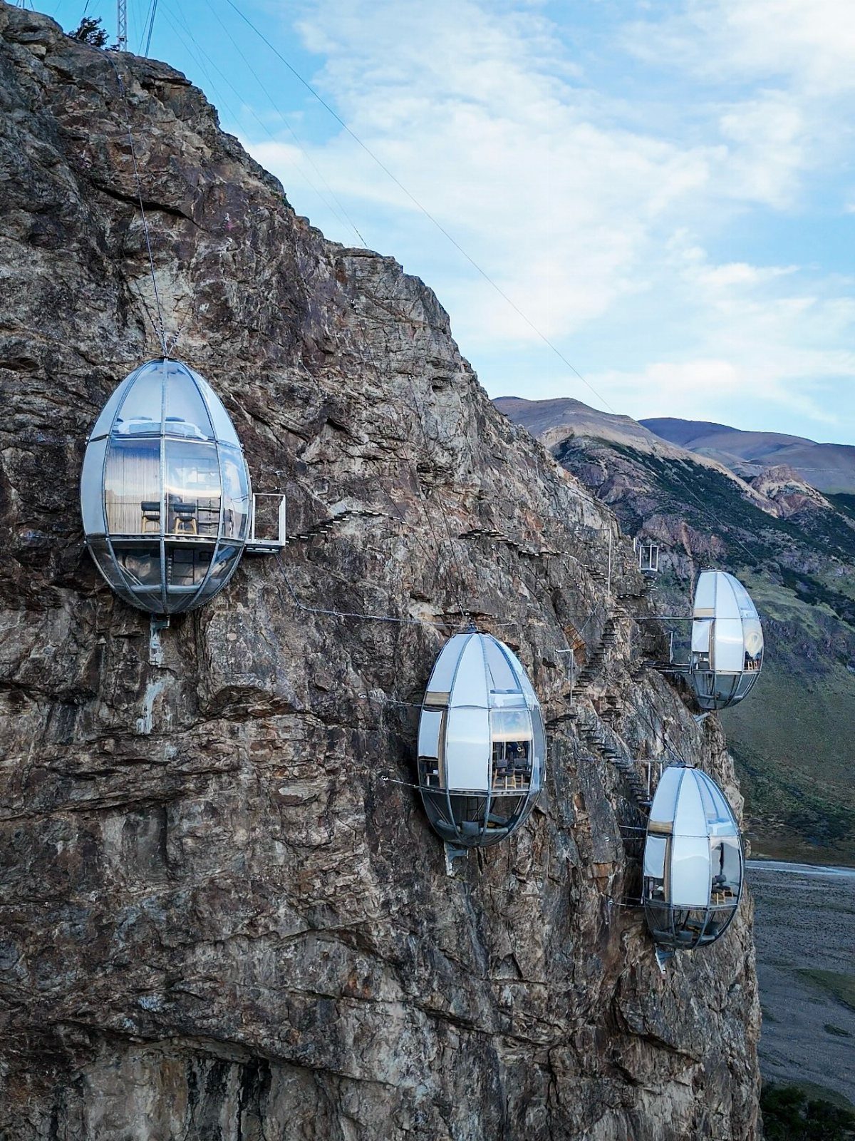 Futuristic glass pods attached to a rocky cliffside with mountains in the background.