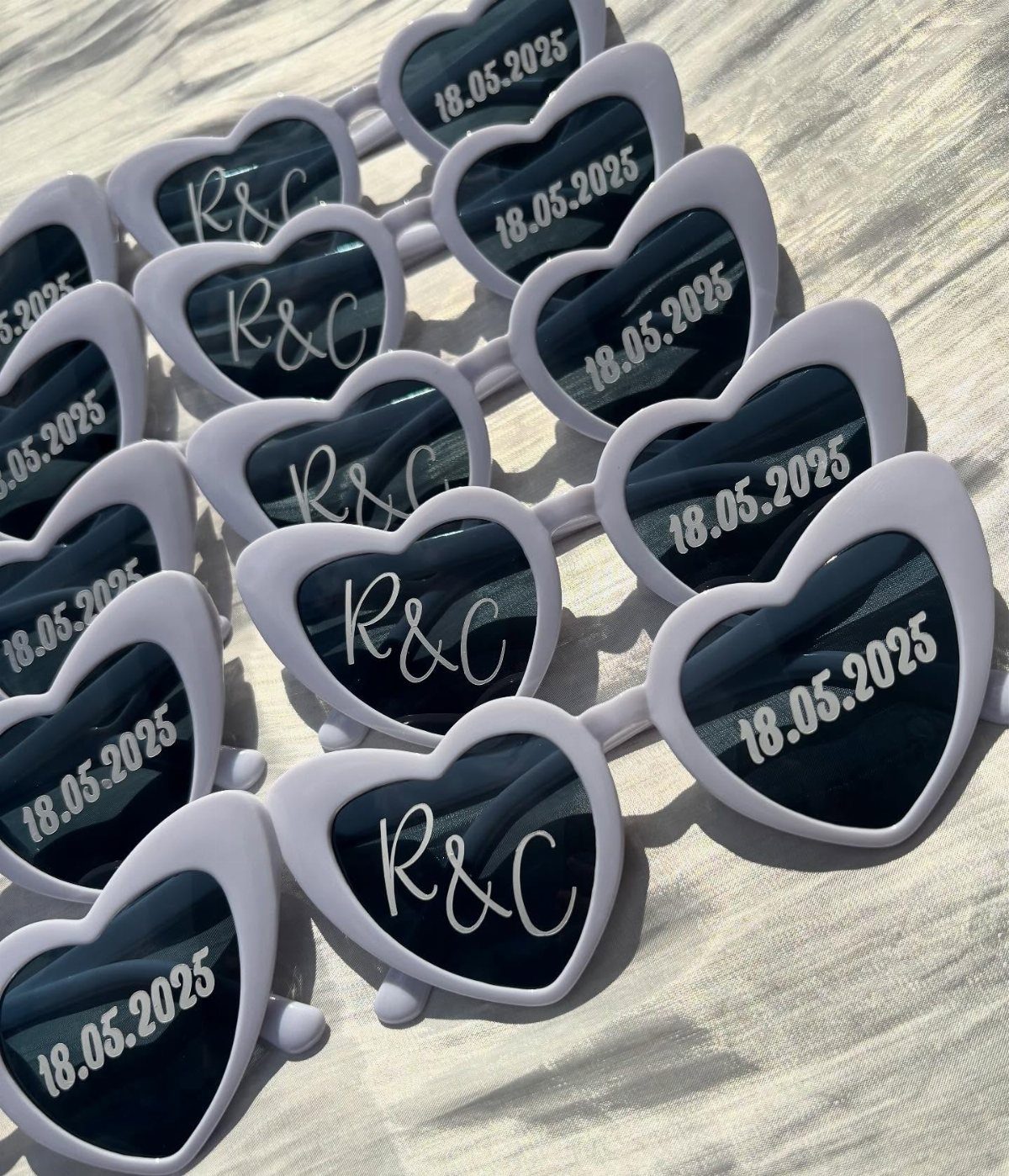 White heart-shaped sunglasses with personalized initials and dates on the dark lenses.