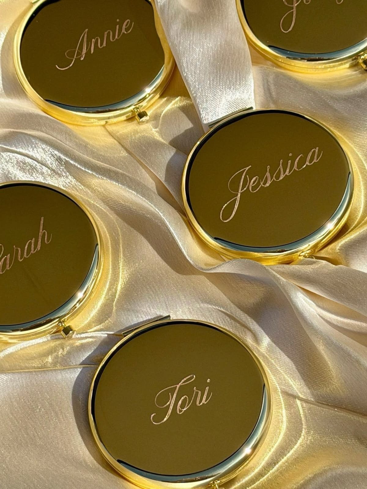 Gold compact mirrors with personalized names resting on champagne satin fabric.