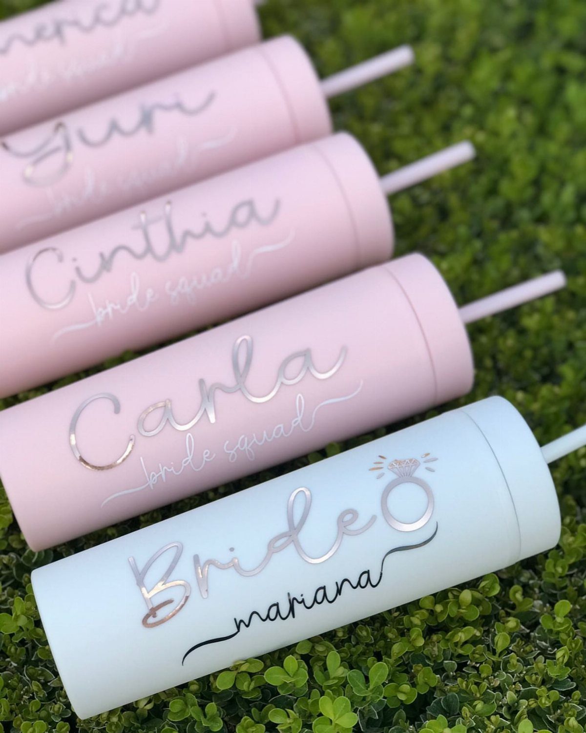 Personalized pink and white tumblers with silver lettering on a green hedge.