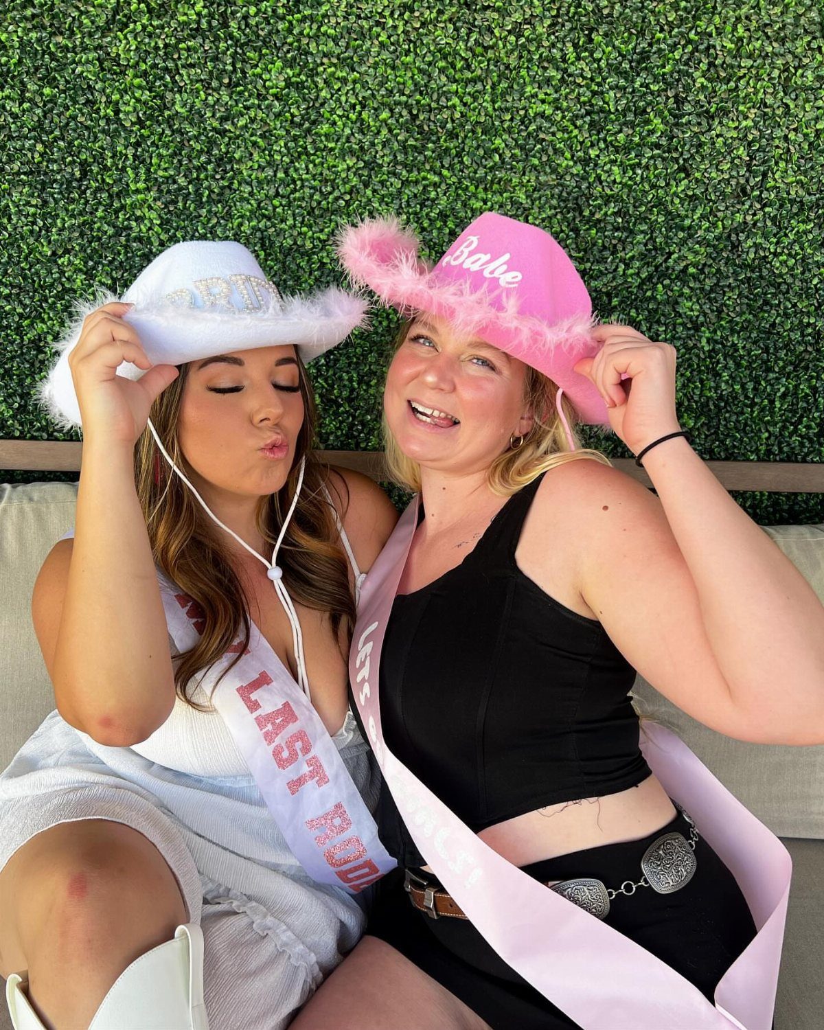 Two women wearing fluffy cowboy hats and sashes posing against a green wall.