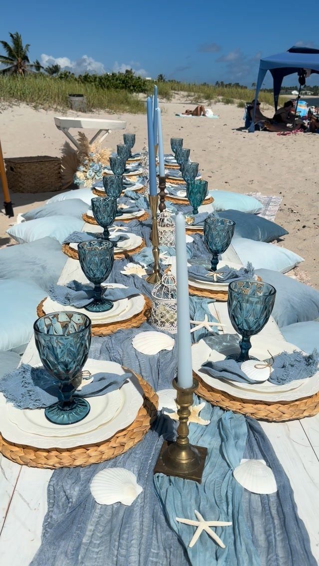 Beach table setting with blue glasses, seashells, and starfish on a sandy shore.
