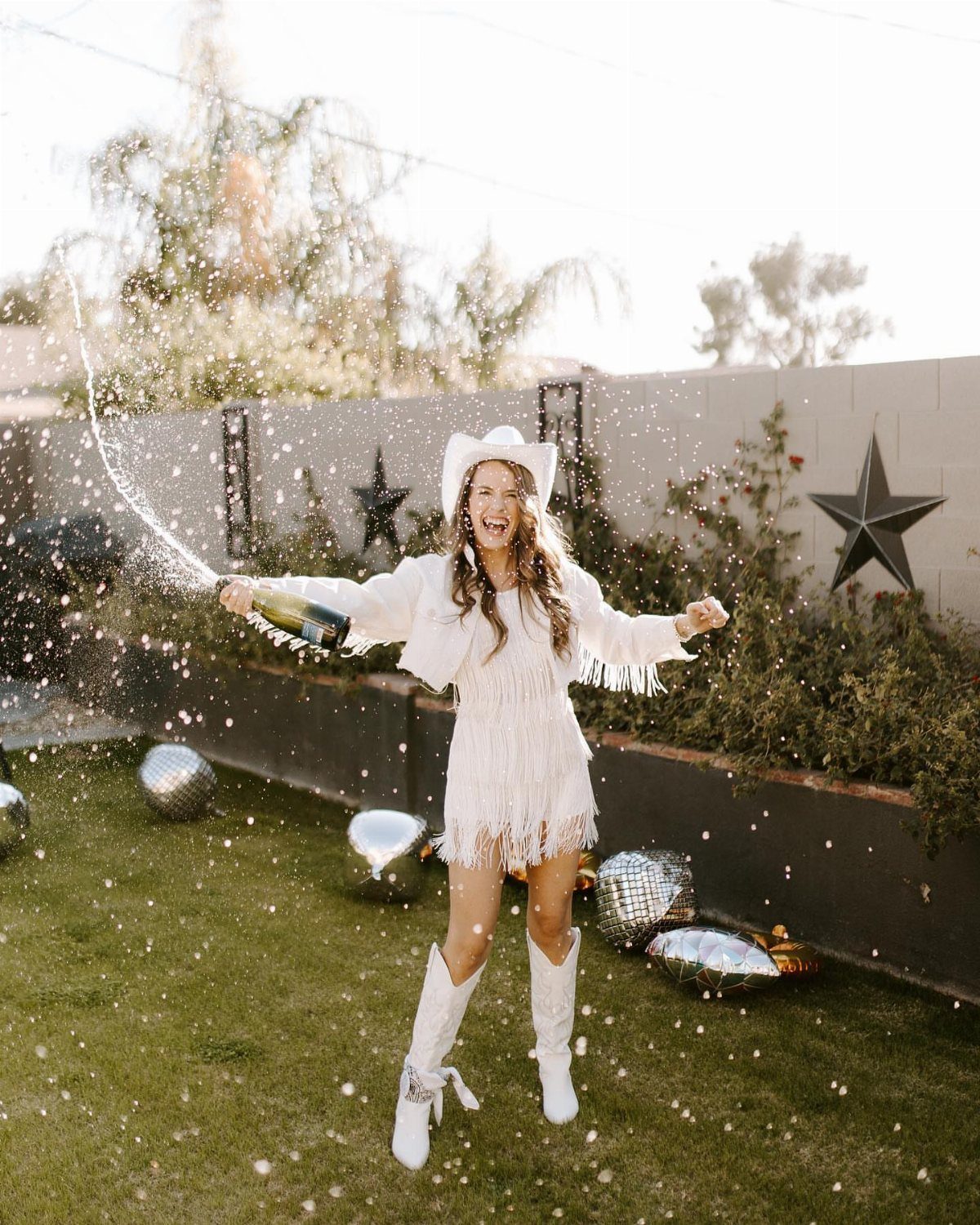 Woman in white western attire popping champagne among silver disco balls on grass