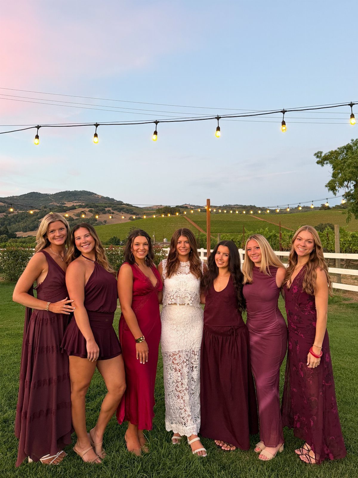 Seven women in burgundy and white dresses posing in a vineyard at sunset.