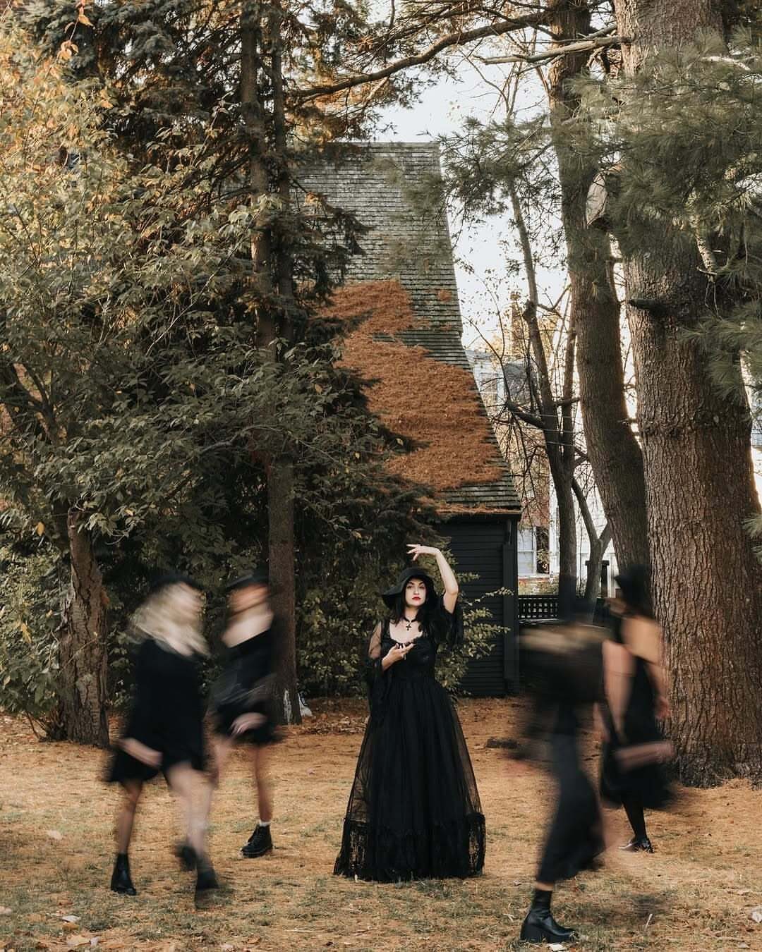 Women in black gothic dresses and hats posing in a wooded autumn setting.
