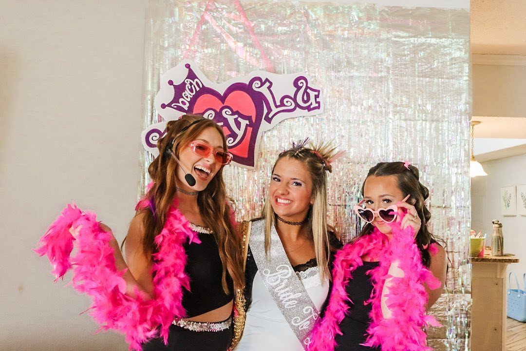 Three women in pink feather boas and heart sunglasses posing for a bachelorette party.