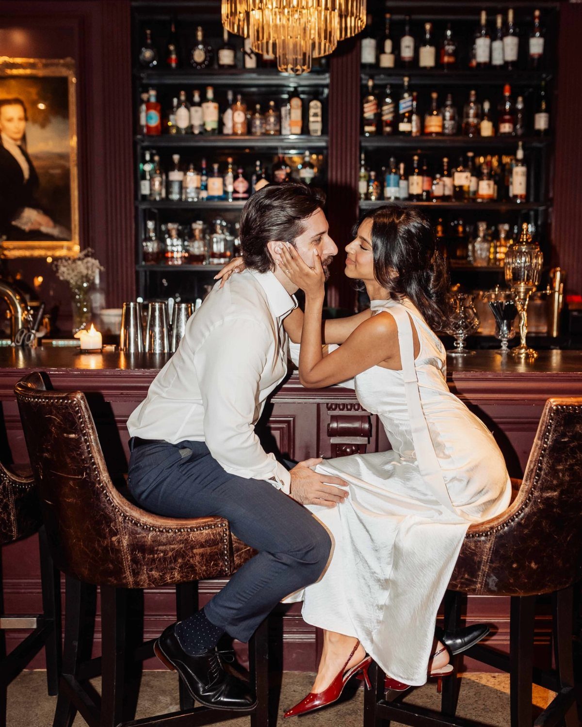 Couple in white formal wear posing romantically on leather stools in a cocktail bar.