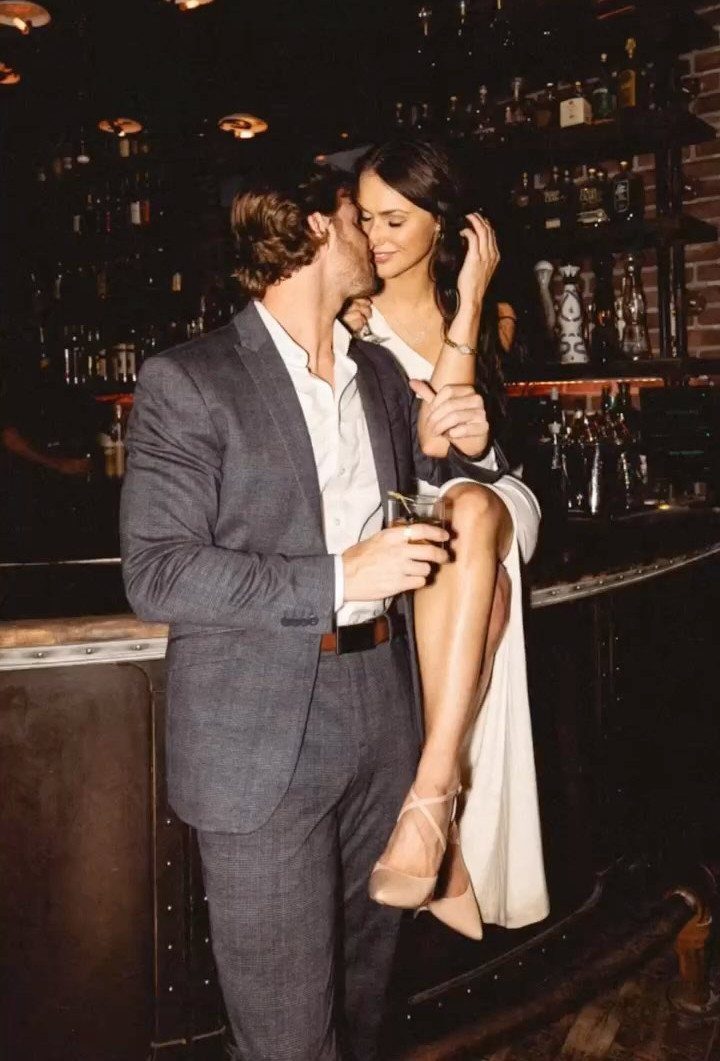 Couple posing intimately at a dark cocktail bar with liquor bottles in background