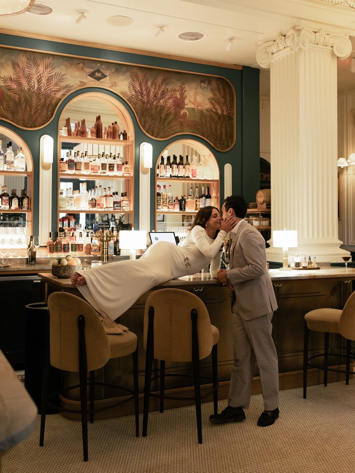 Couple in formal wear posing creatively at an elegant bar with liquor shelves.