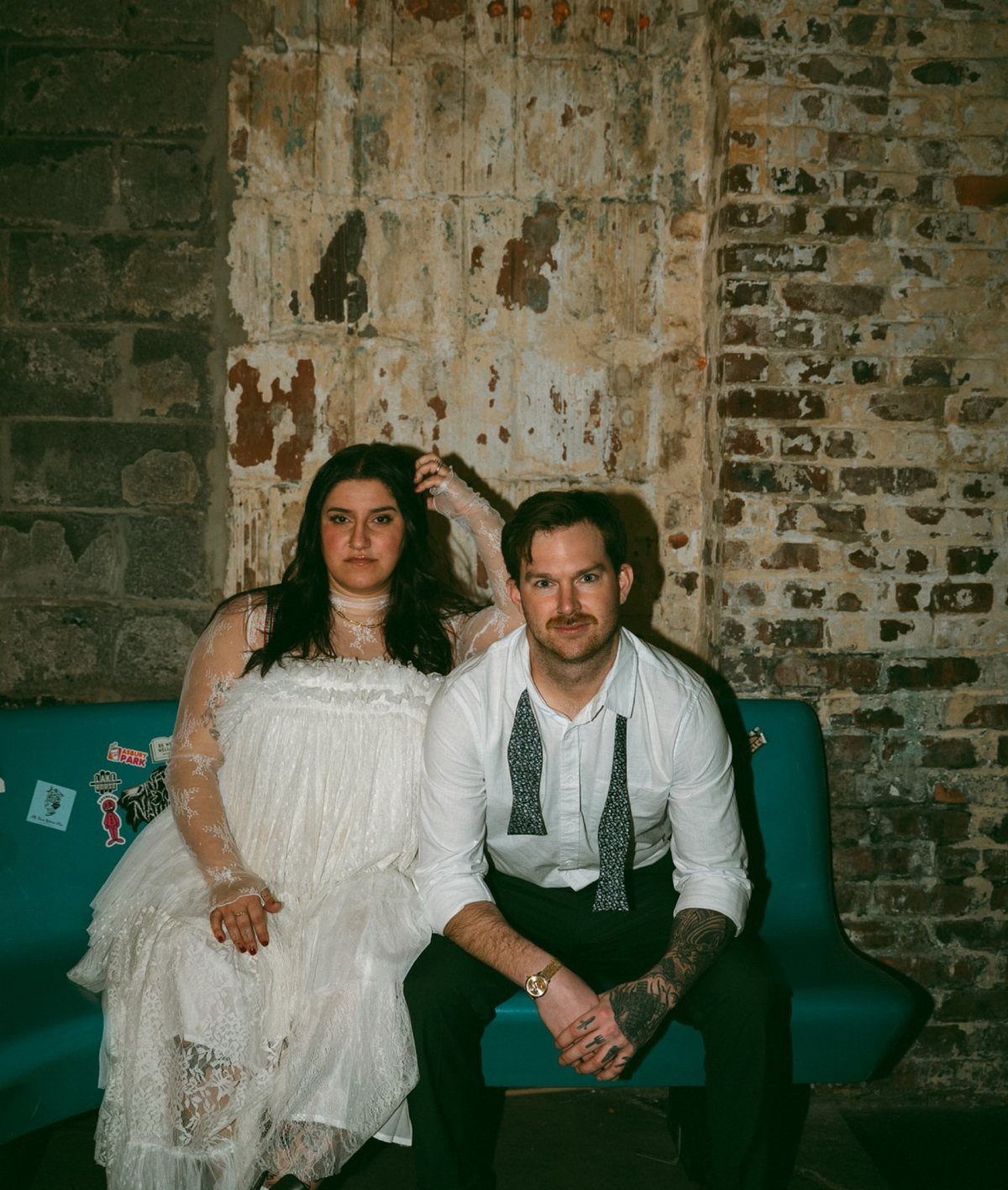 Couple sitting on a teal couch against a brick wall with high-contrast flash lighting.