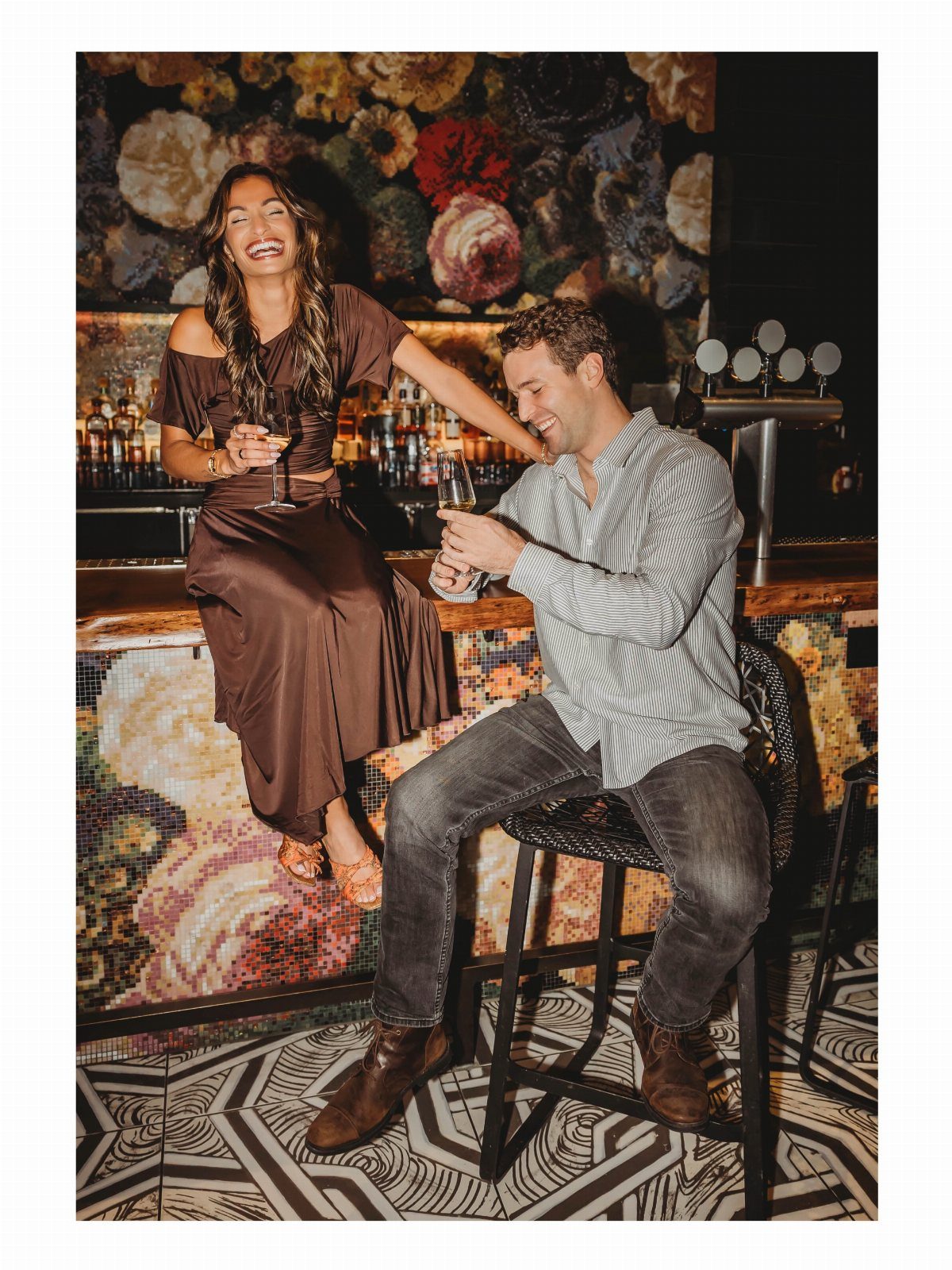 Smiling couple with wine glasses posing at a bar with floral decor