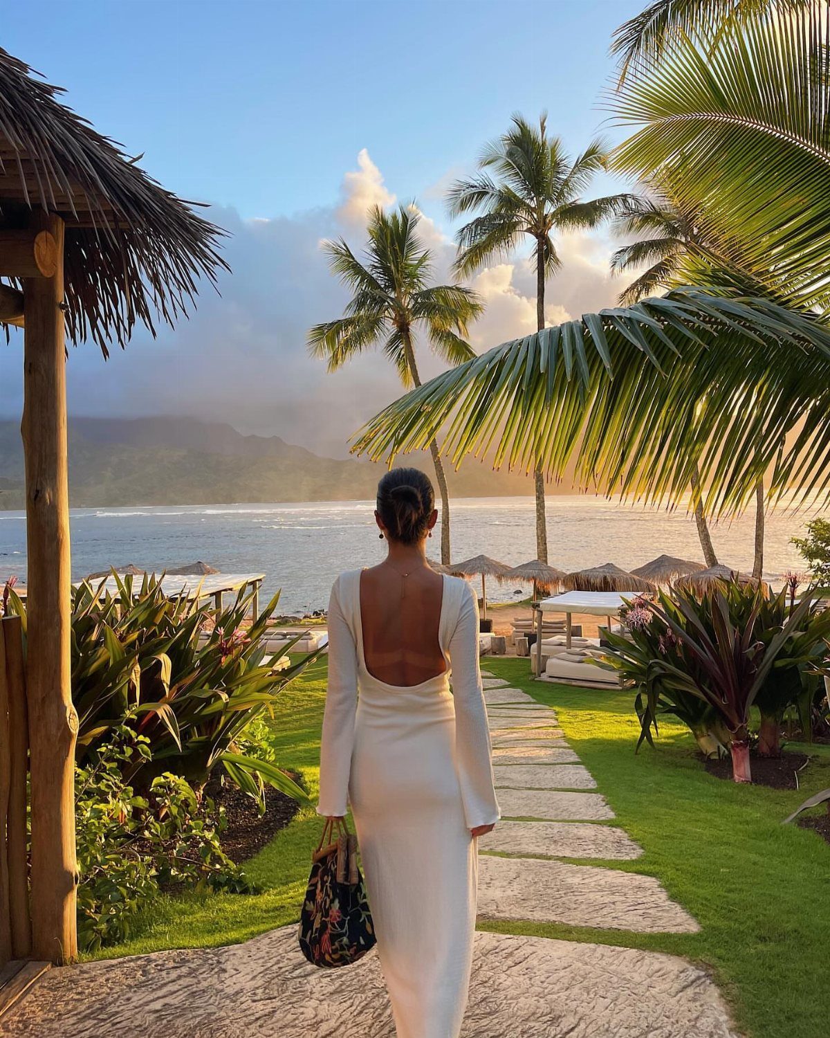 Woman in a long white open-back dress walking toward a tropical beach