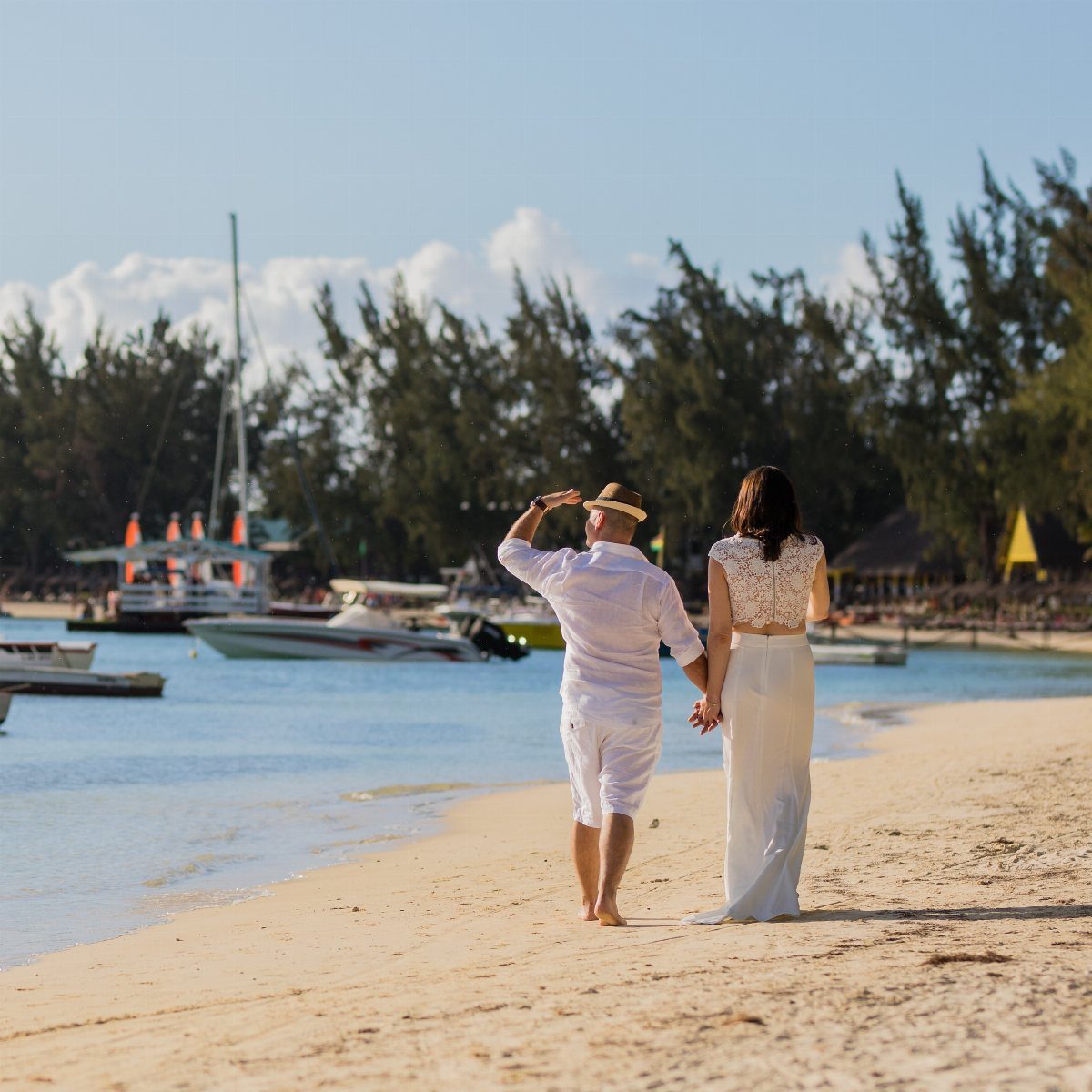 Couple in matching white beach outfits walking hand-in-hand along a sunny shoreline.