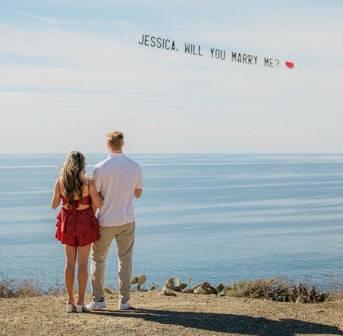 Couple standing on a cliff looking at a proposal banner in the sky.