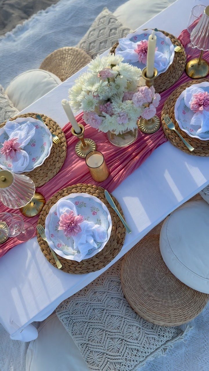 Romantic beach picnic setup with a low white table, pink runner, and floral arrangements.