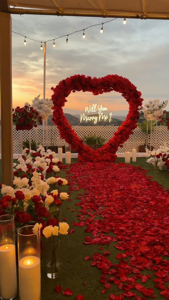 Heart-shaped red rose arch and petal carpet at sunset for a romantic proposal.