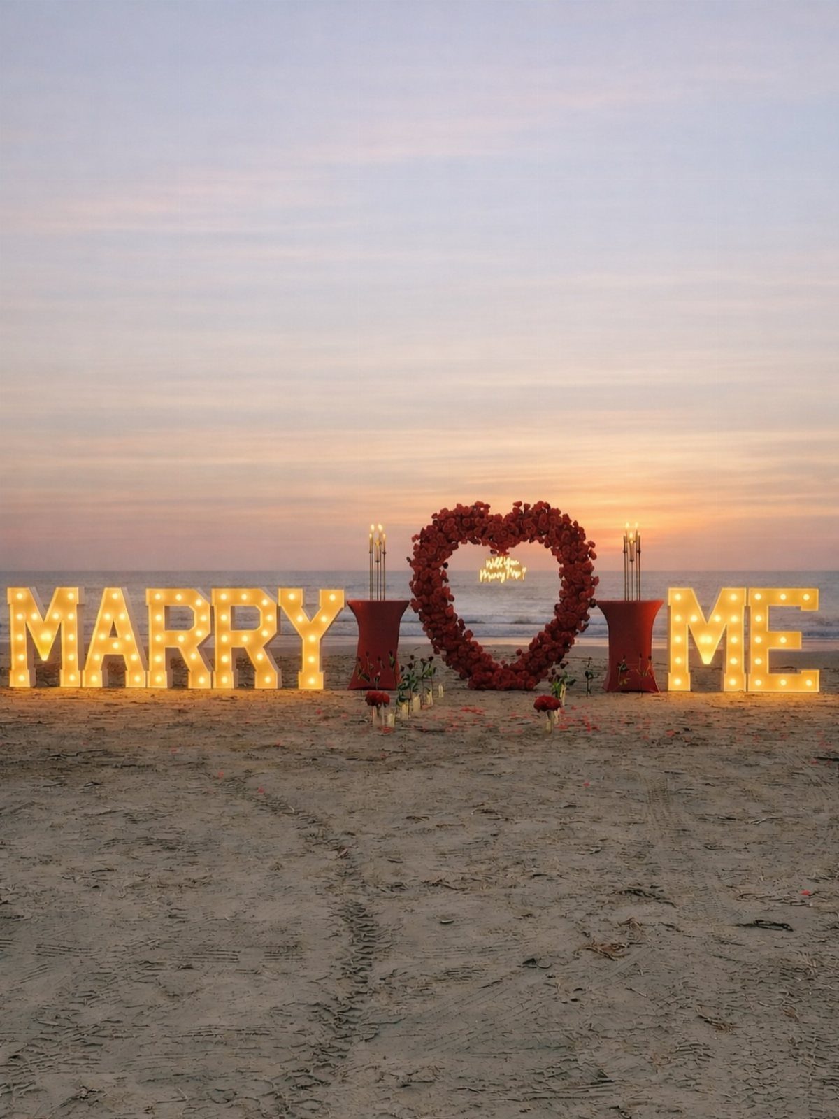 Illuminated Marry Me letters and a red rose heart on a beach sunset.
