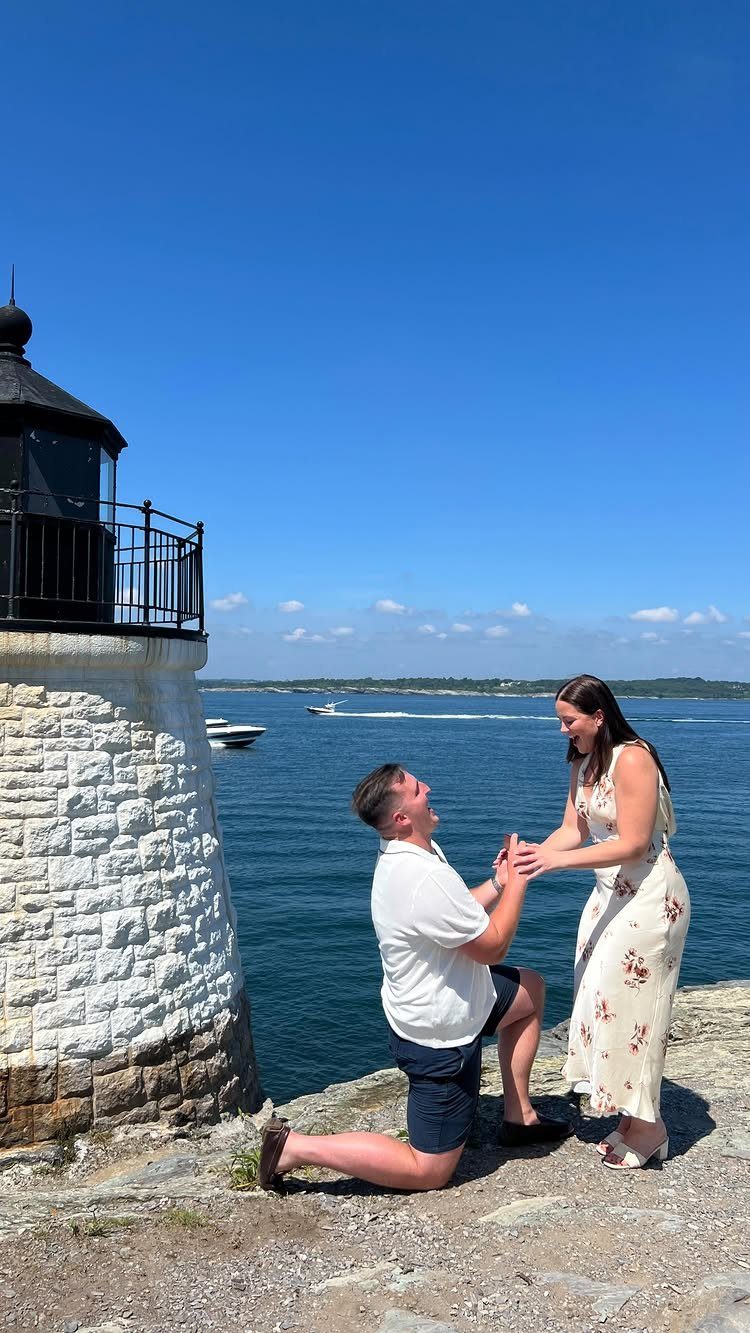 Man kneeling to propose to a woman on a rocky coast by a lighthouse.