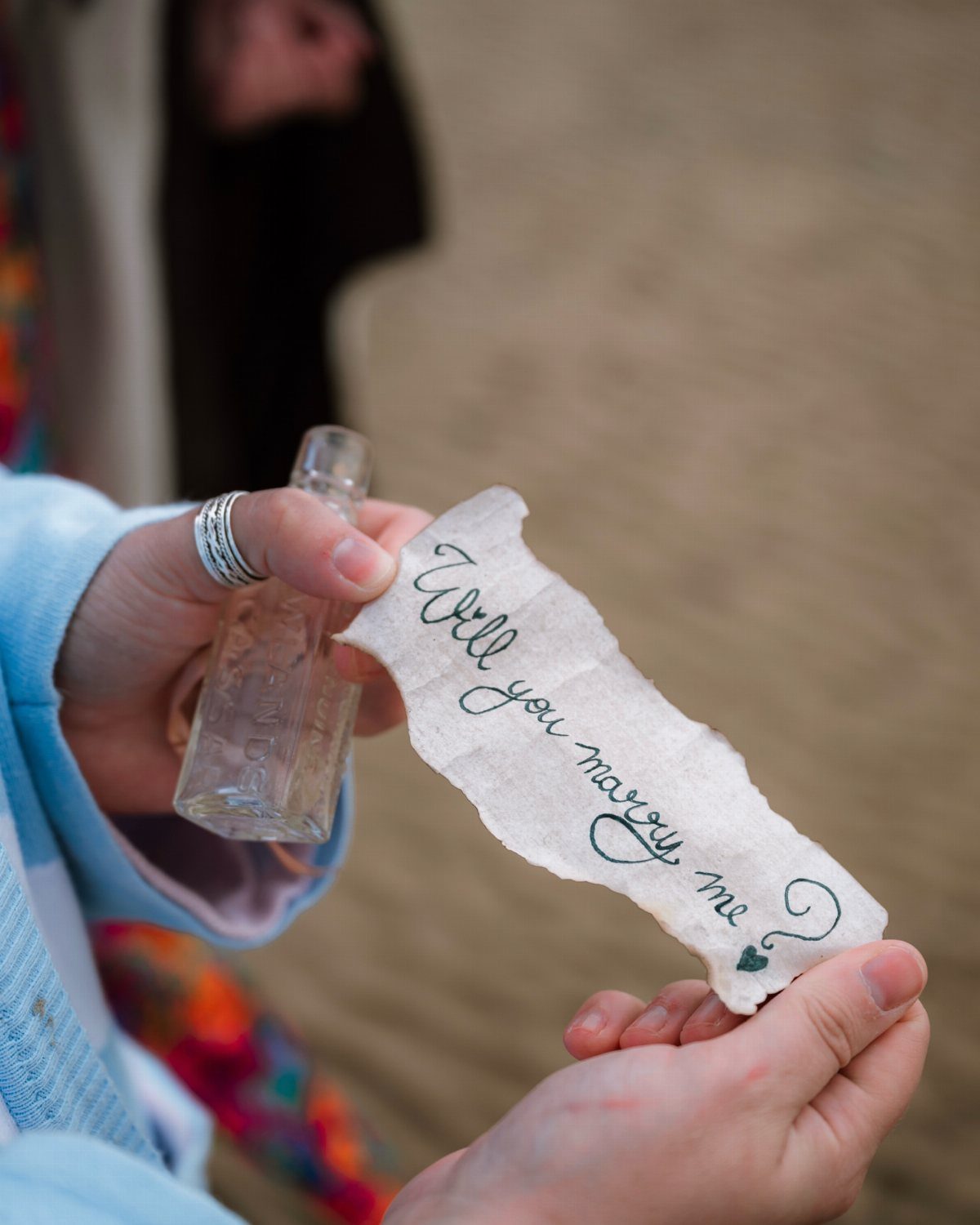 Hands holding a glass bottle and a proposal note on a sandy beach.