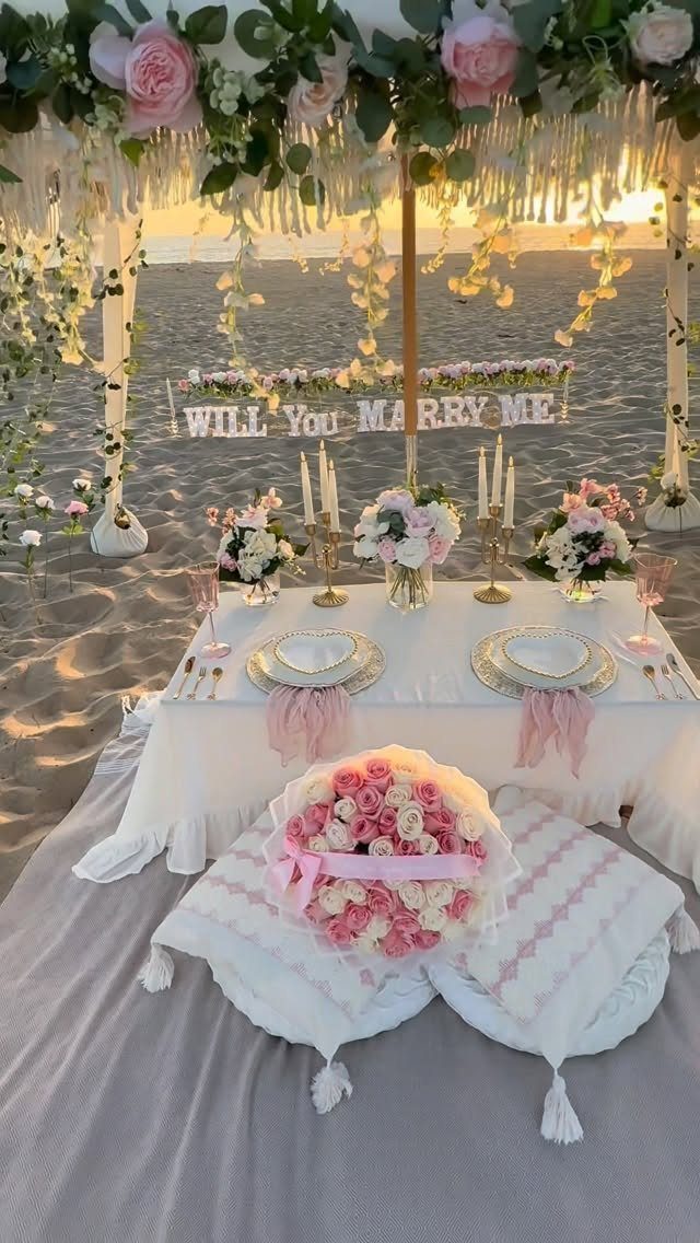 Romantic beach proposal setup with a dinner table, floral canopy, and marriage proposal sign.