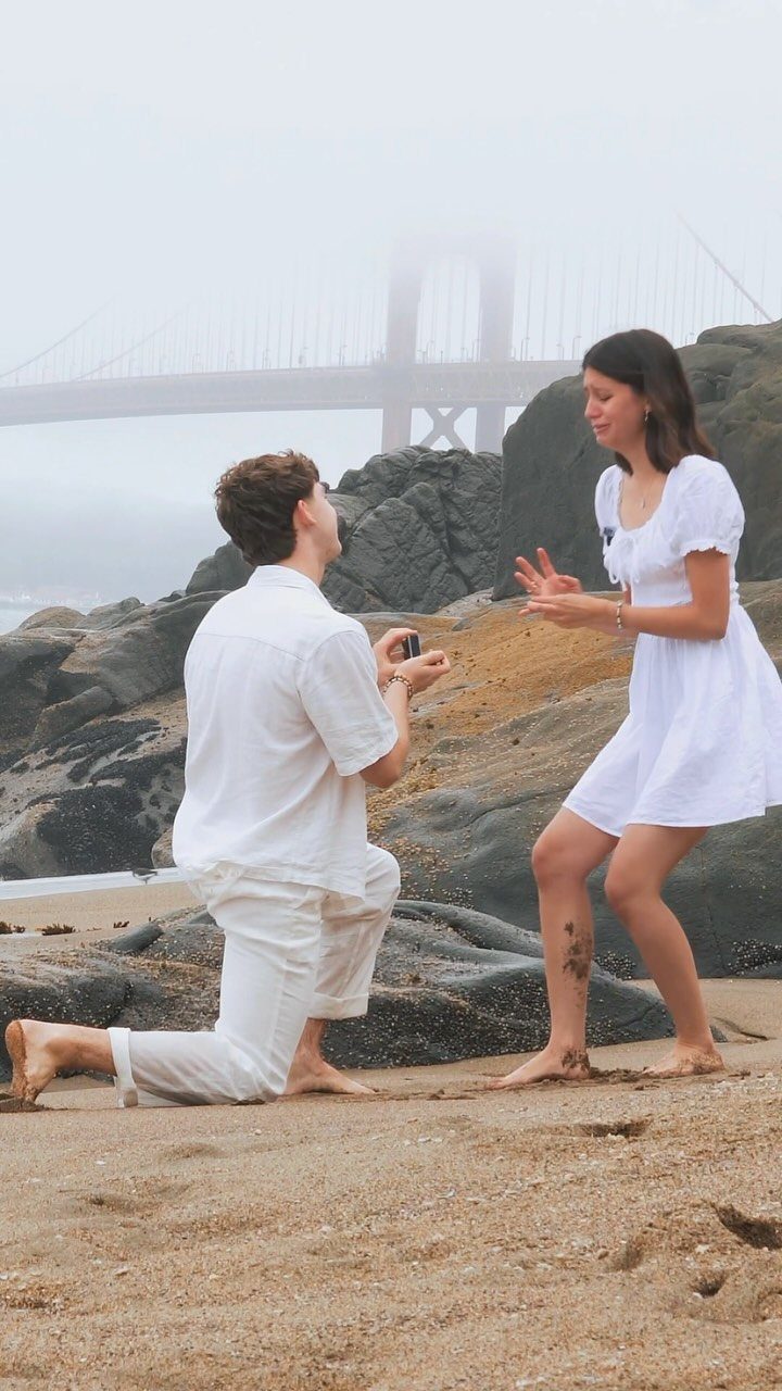 Man kneeling on a sandy beach proposing to a woman with a bridge background