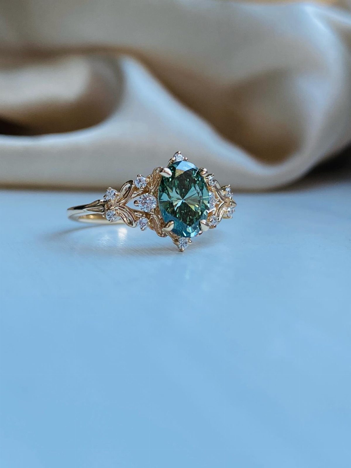 Gold engagement ring with teal gemstone and intricate leaf and vine engravings.