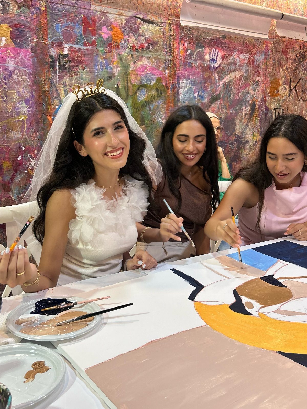 Three women painting a large abstract canvas in a colorful art studio.