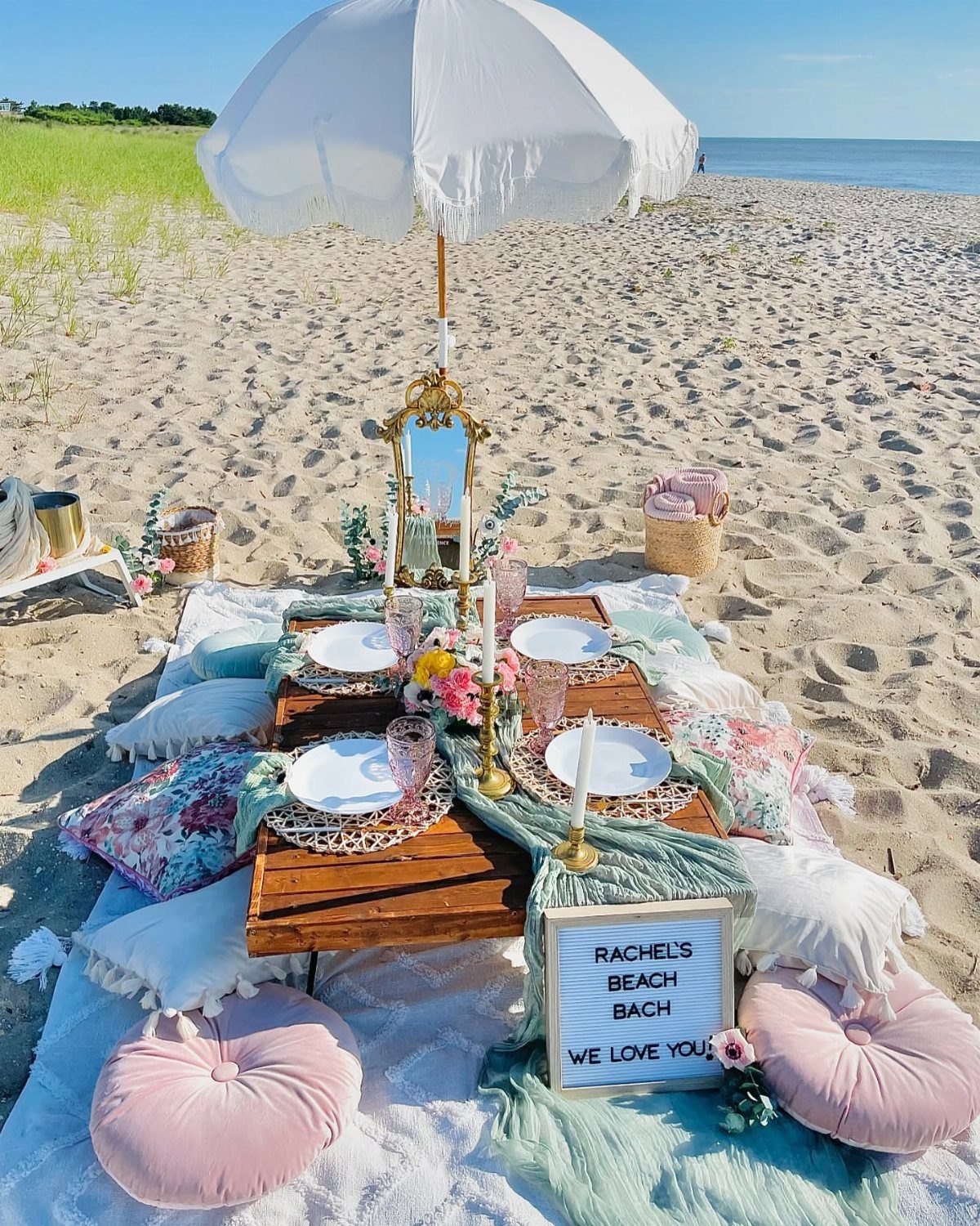 Beach picnic setup with a low wooden table, white umbrella, and pink cushions.