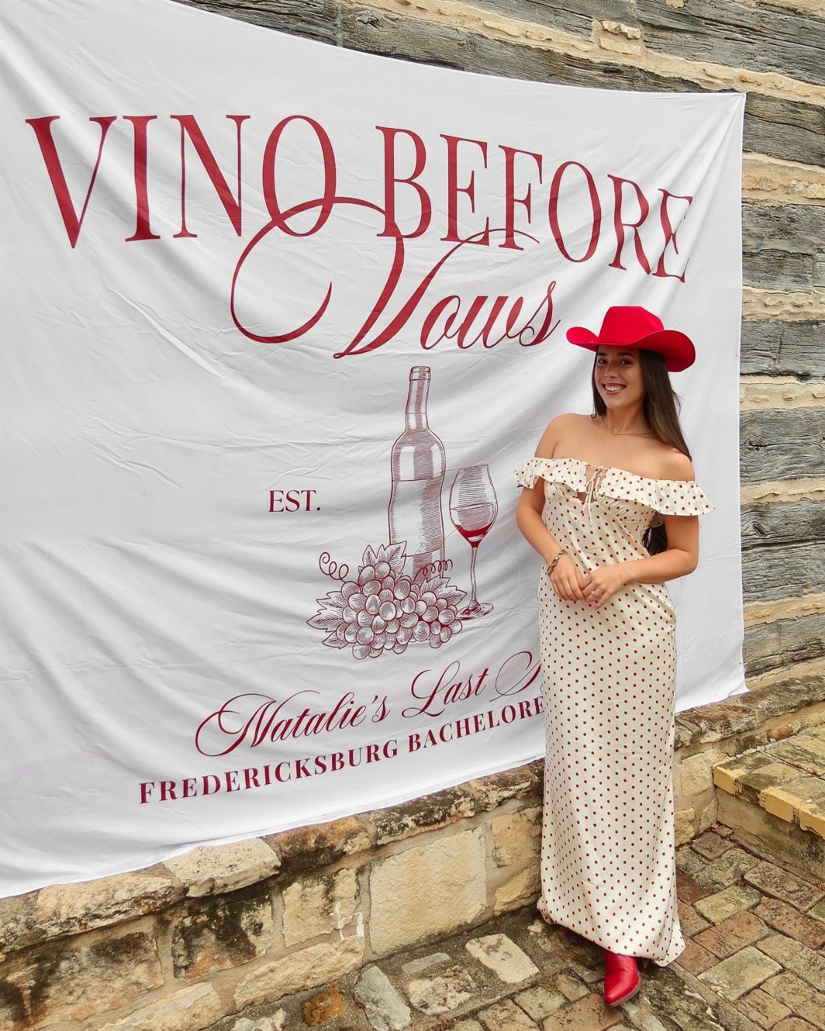 Woman in red hat and polka-dot dress posing by a wine-themed bachelorette banner.
