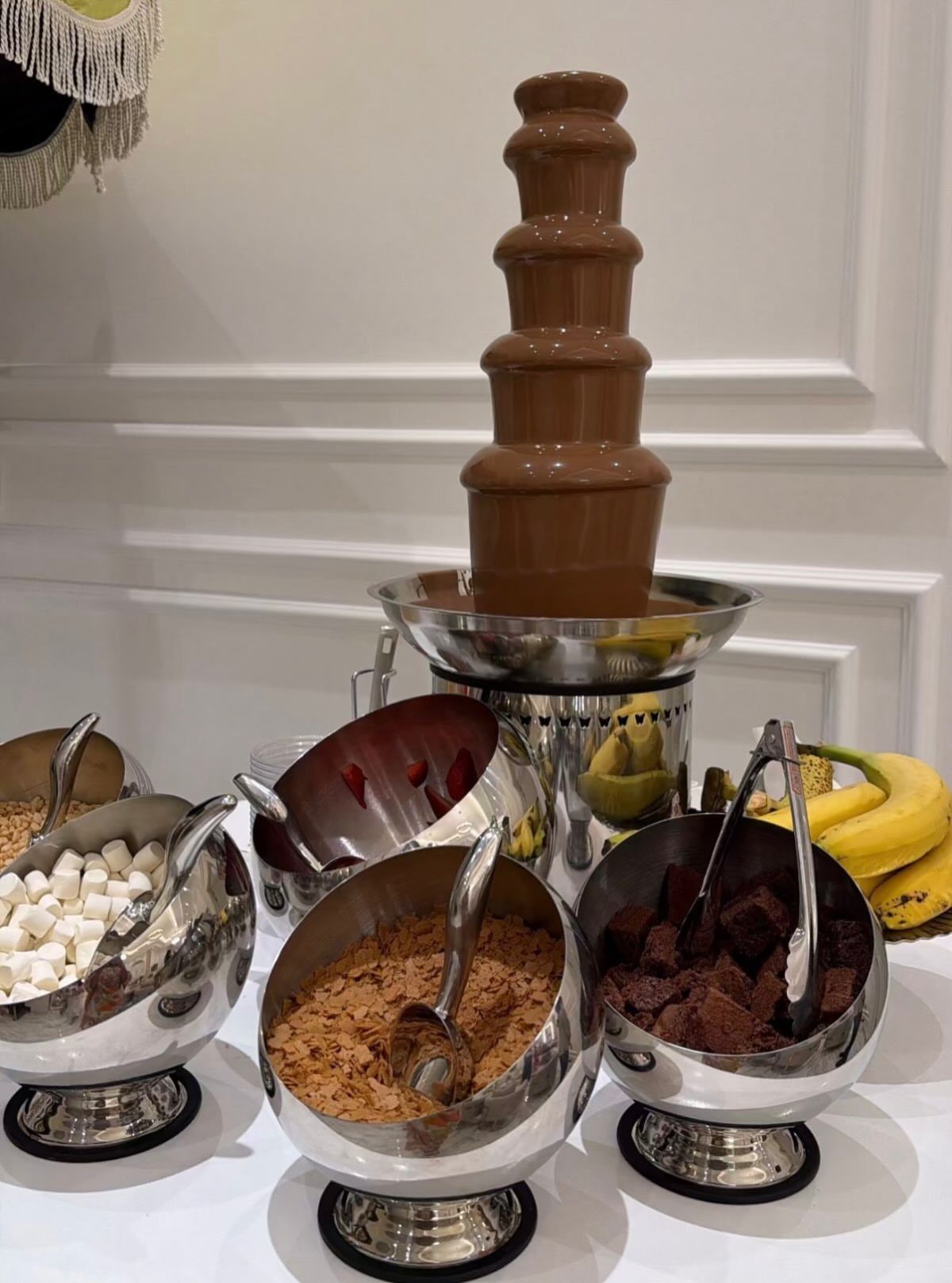 Chocolate fountain with tiered flowing chocolate and bowls of dipping treats on white table