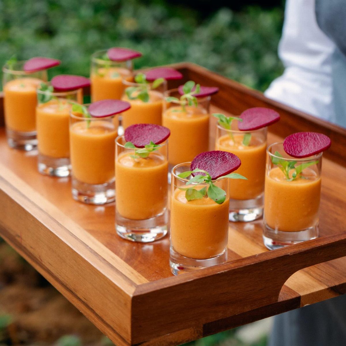 Orange gazpacho shots garnished with red radish slices on a wooden tray