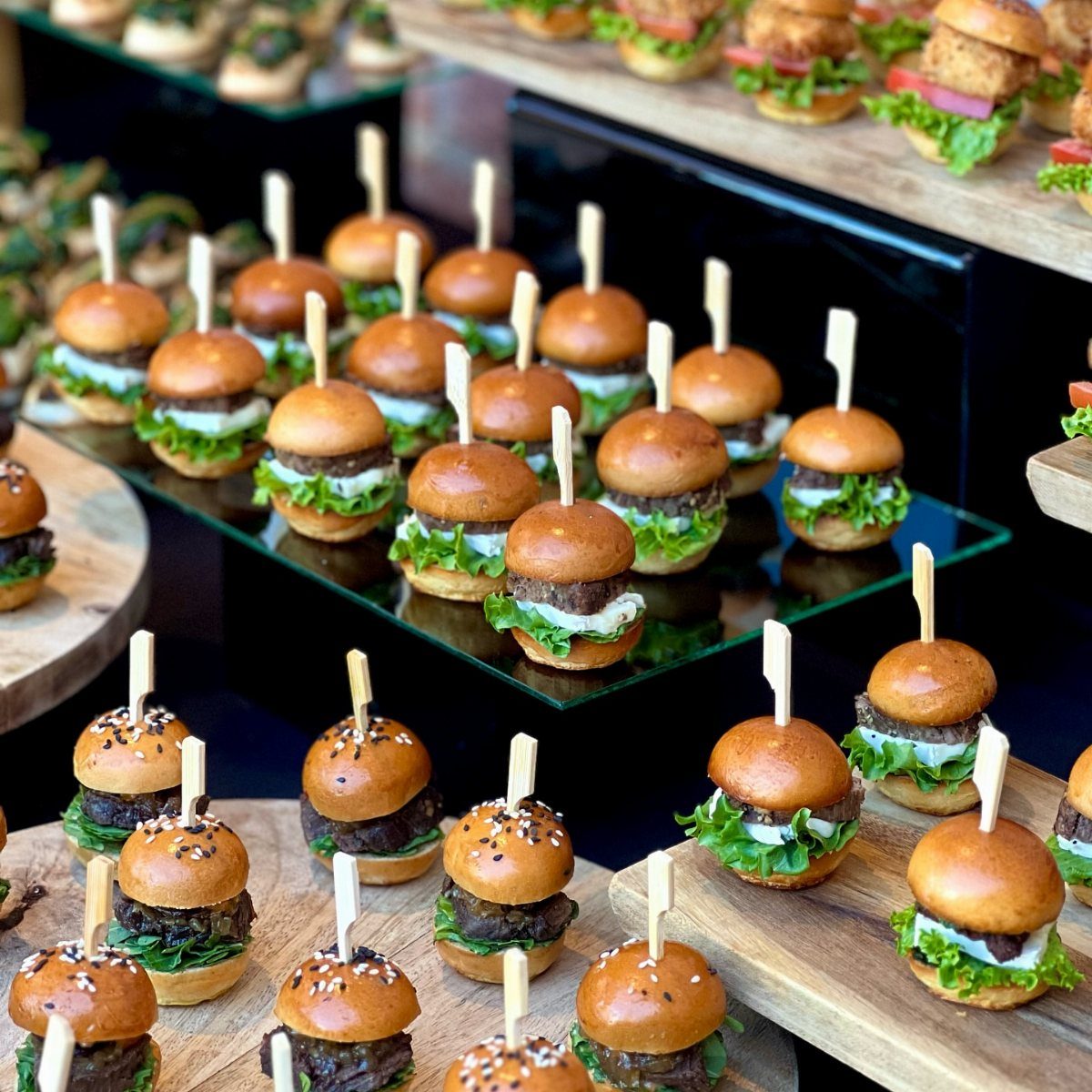 Mini beef sliders with cheese and lettuce on wooden and glass platters.