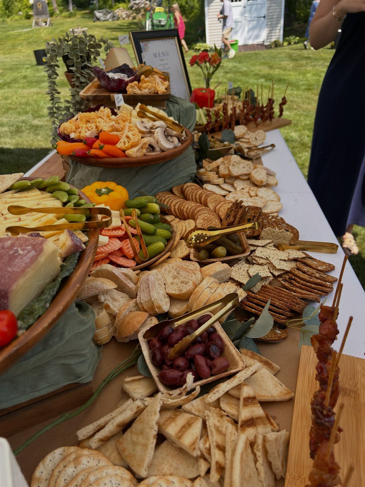 Lavish grazing table with cheeses, crackers, olives, and shrimp outdoors on white cloth.