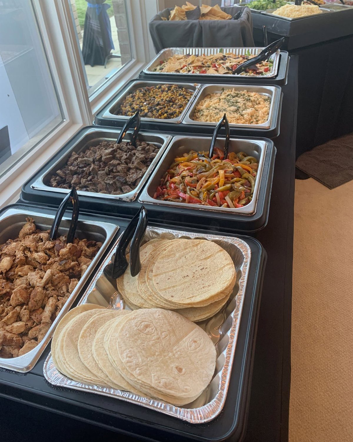 Taco bar buffet with meat, vegetables, rice, and tortillas in metal trays.