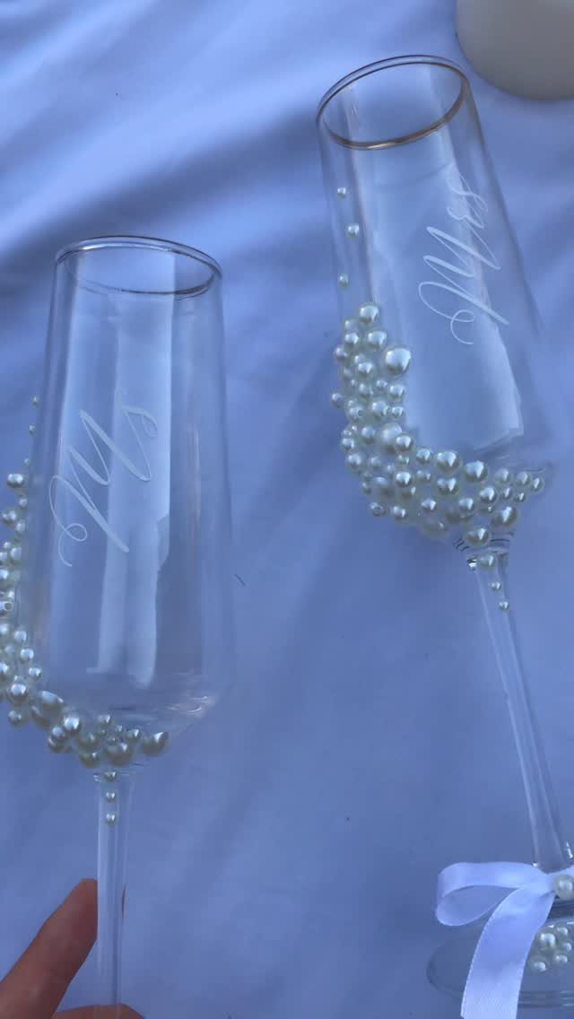 Two champagne flutes with etched Mrs. text and white pearl decorations on white fabric