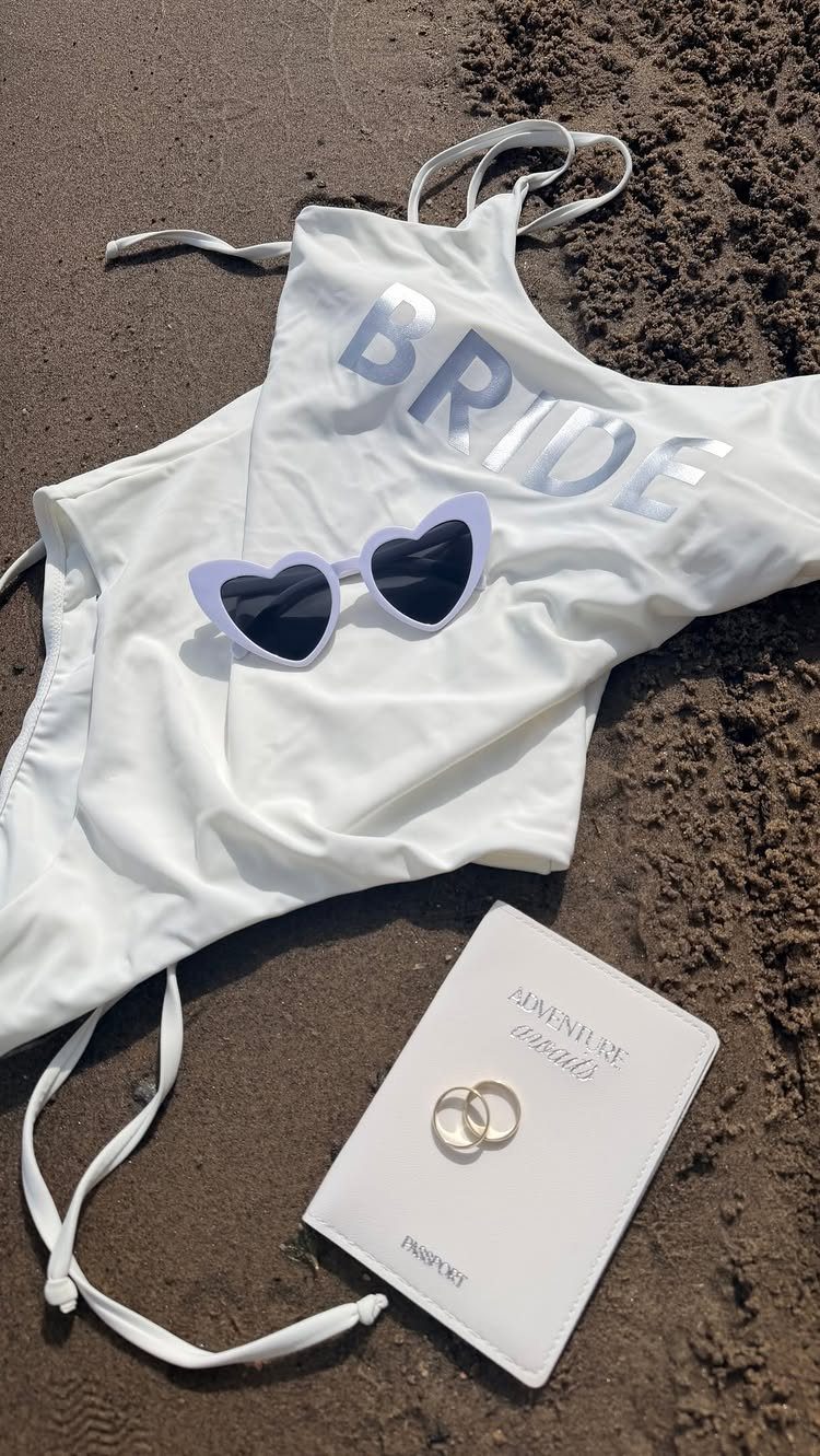White bride swimsuit, heart sunglasses, wedding rings, and passport cover on beach sand.