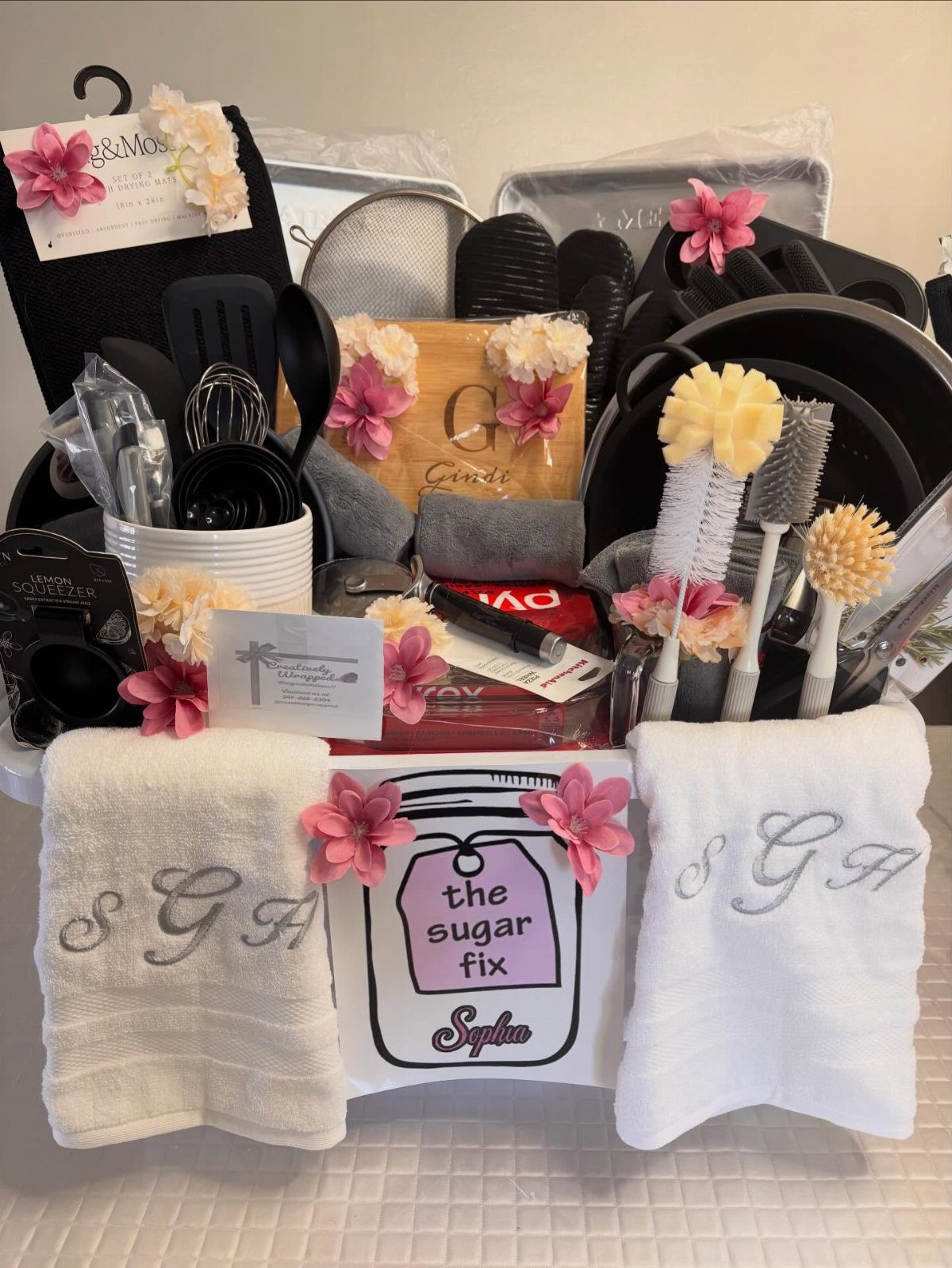 Gift basket with monogrammed towels, kitchen utensils, and cookware decorated with pink flowers.
