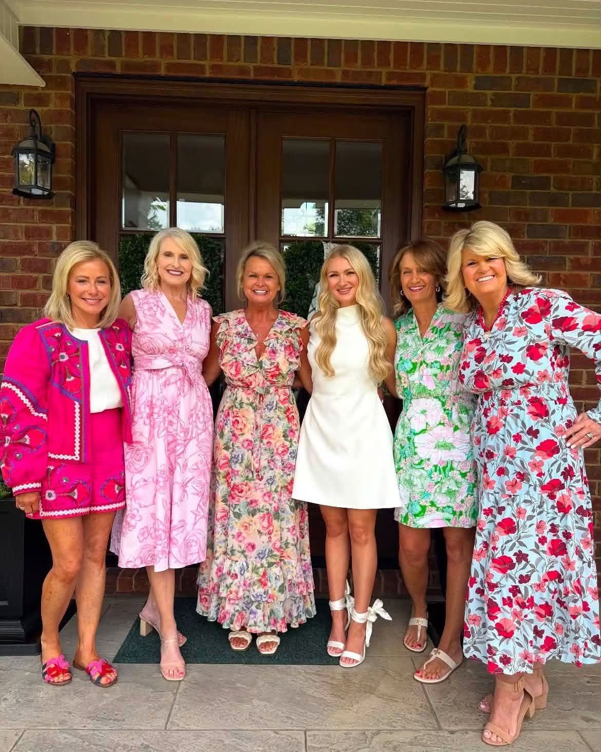 Six women posing in colorful floral dresses and outfits in front of a house.