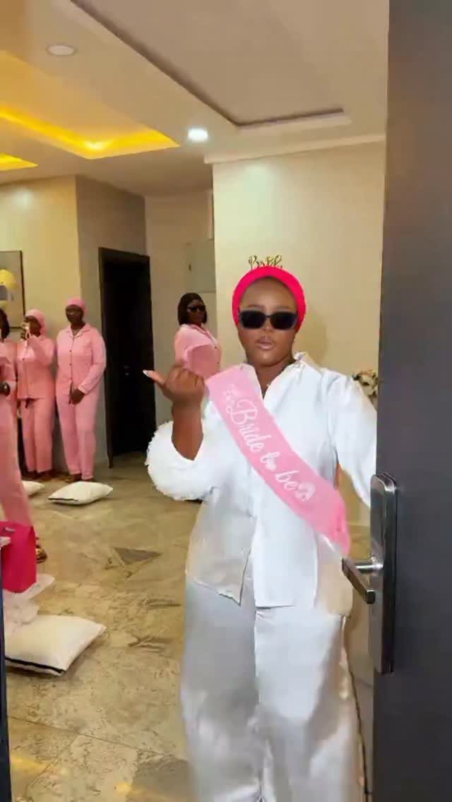 Woman in white satin pajamas and pink bride-to-be sash posing indoors with friends.