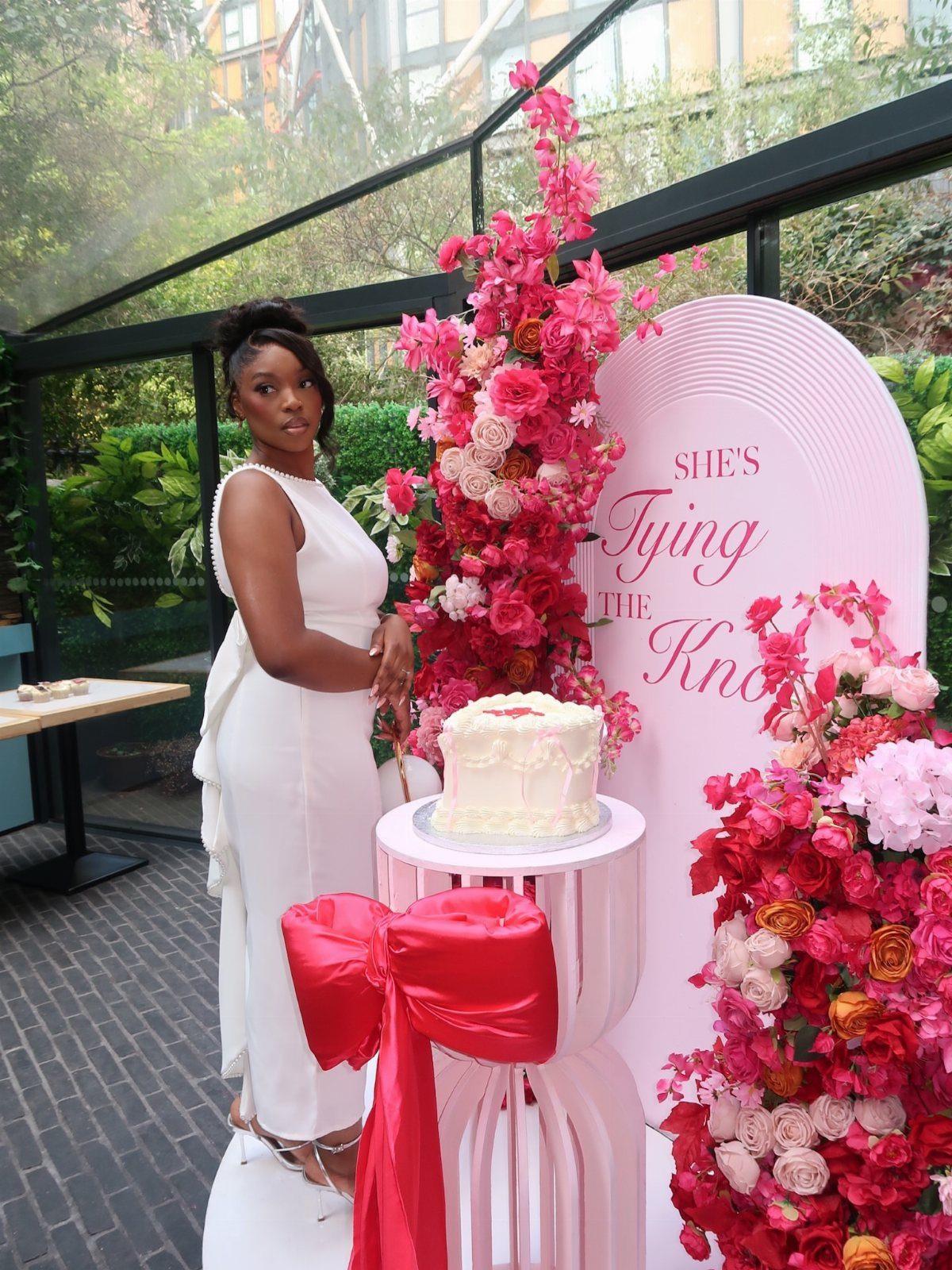 Woman in white midi dress standing by a cake and pink floral decorations.