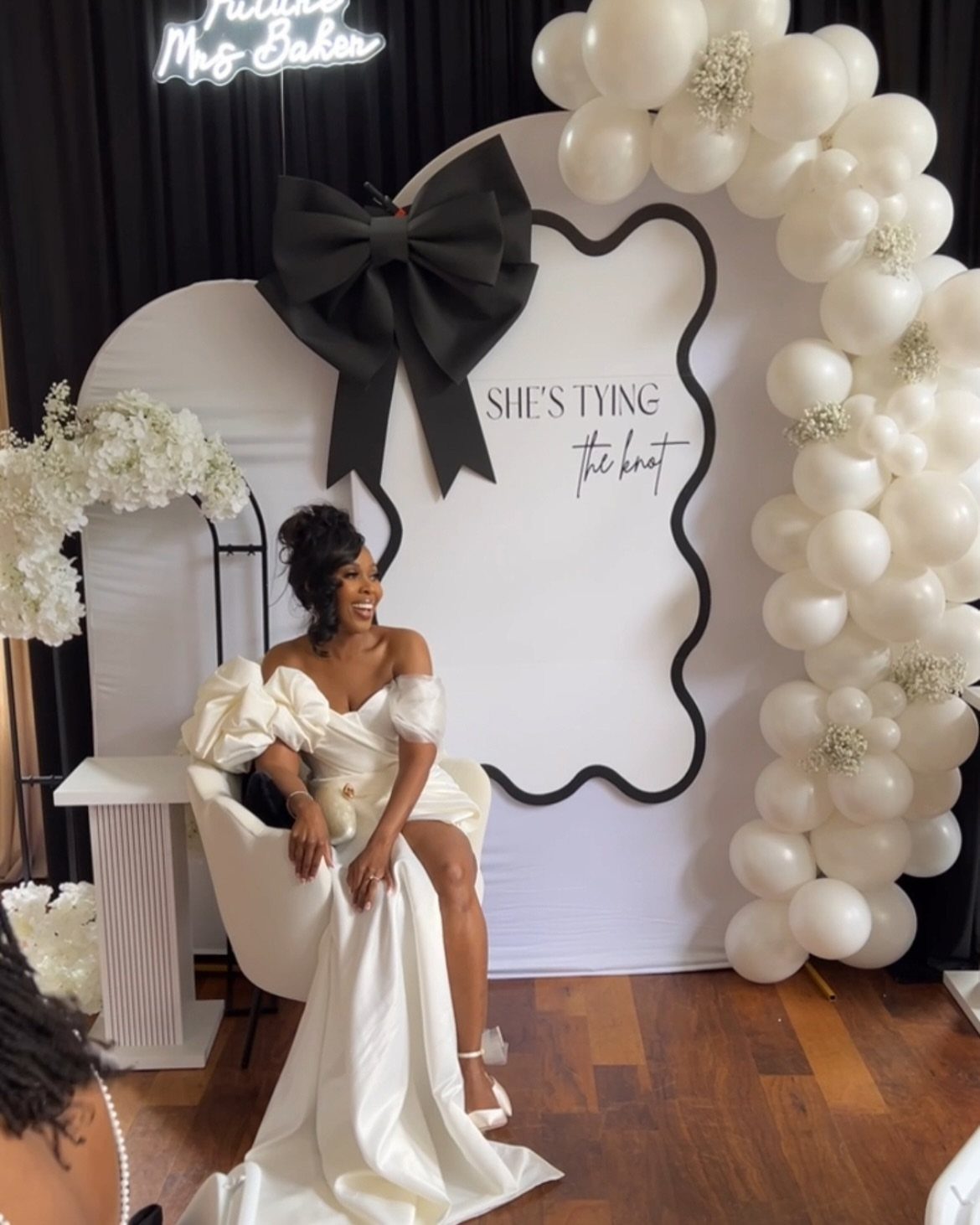 Woman in white satin off-the-shoulder gown sitting before bridal shower balloon backdrop