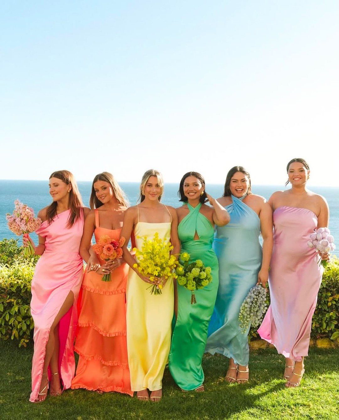 Six women wearing vibrant, colorful satin maxi dresses standing outdoors by the ocean.