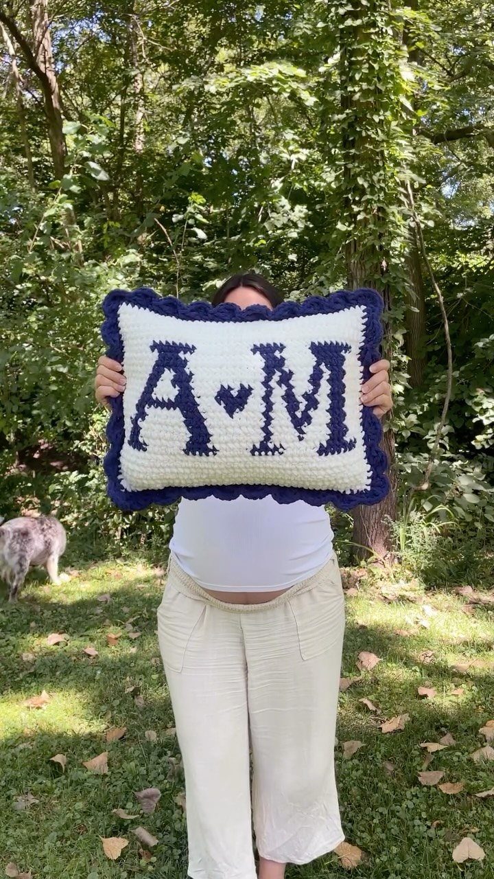Crocheted white throw pillow with navy blue initials and heart held outdoors.