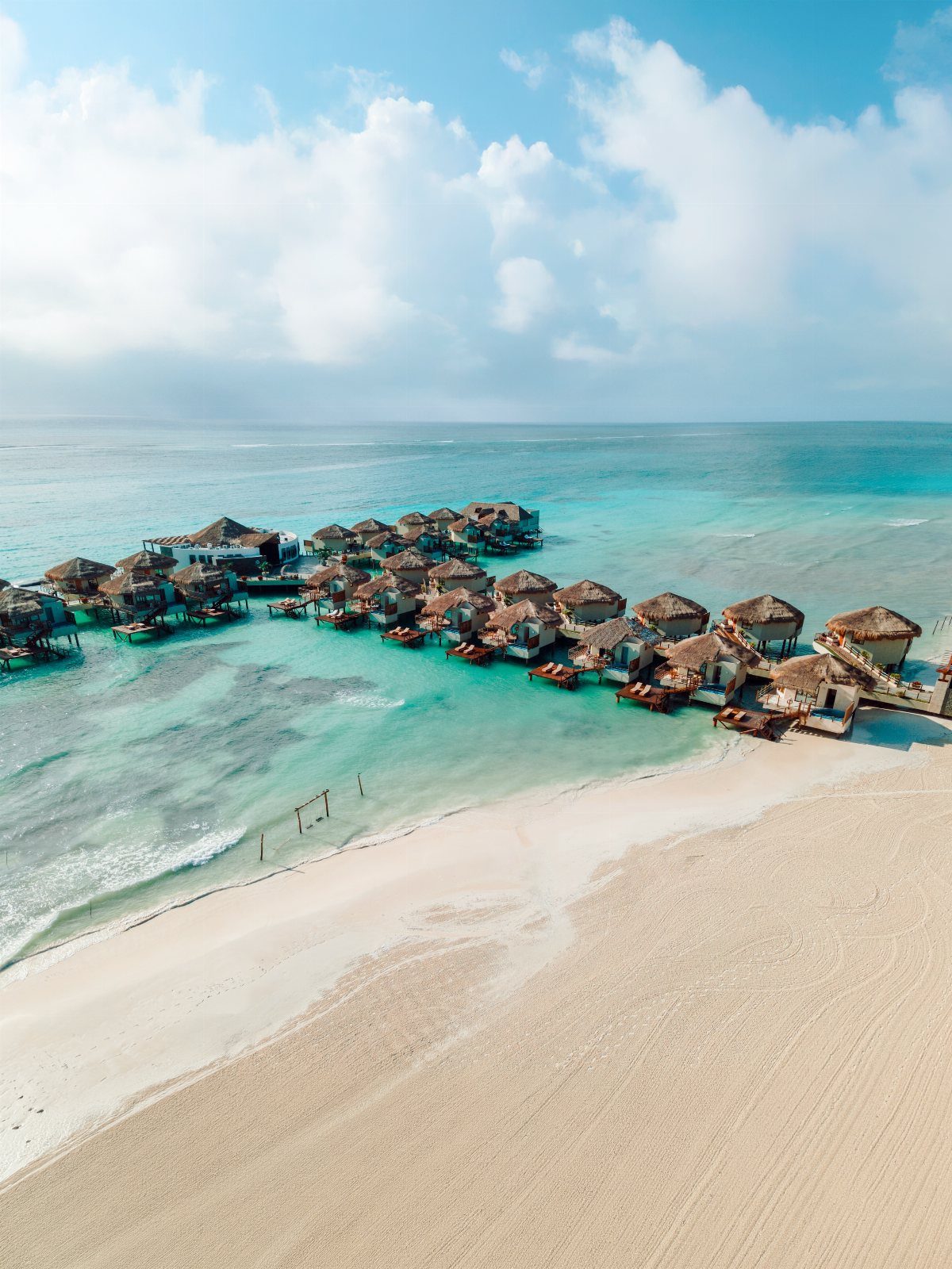 Overwater bungalows with thatched roofs on a white sand beach in turquoise water.